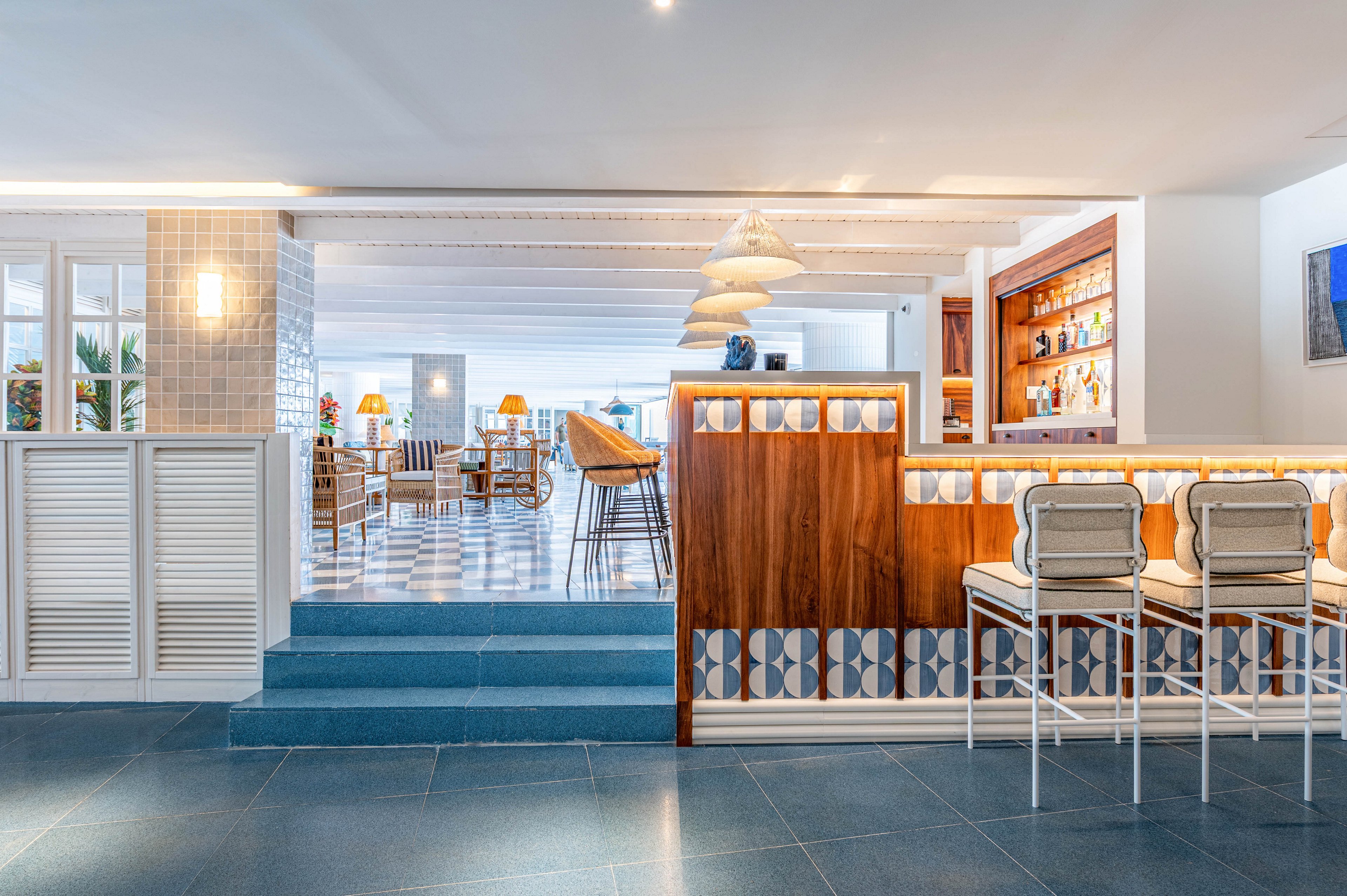 Spacious restaurant interior features a wooden bar with cushioned stools in the foreground. The setting is bright and airy, with blue-tiled floors, white walls, and patterned accents creating a coastal atmosphere. Pendant lights hang above the bar, while additional tables and chairs extend into the background dining area. Warm lighting from lamps and the bar shelves adds a welcoming, contemporary feel to the space.