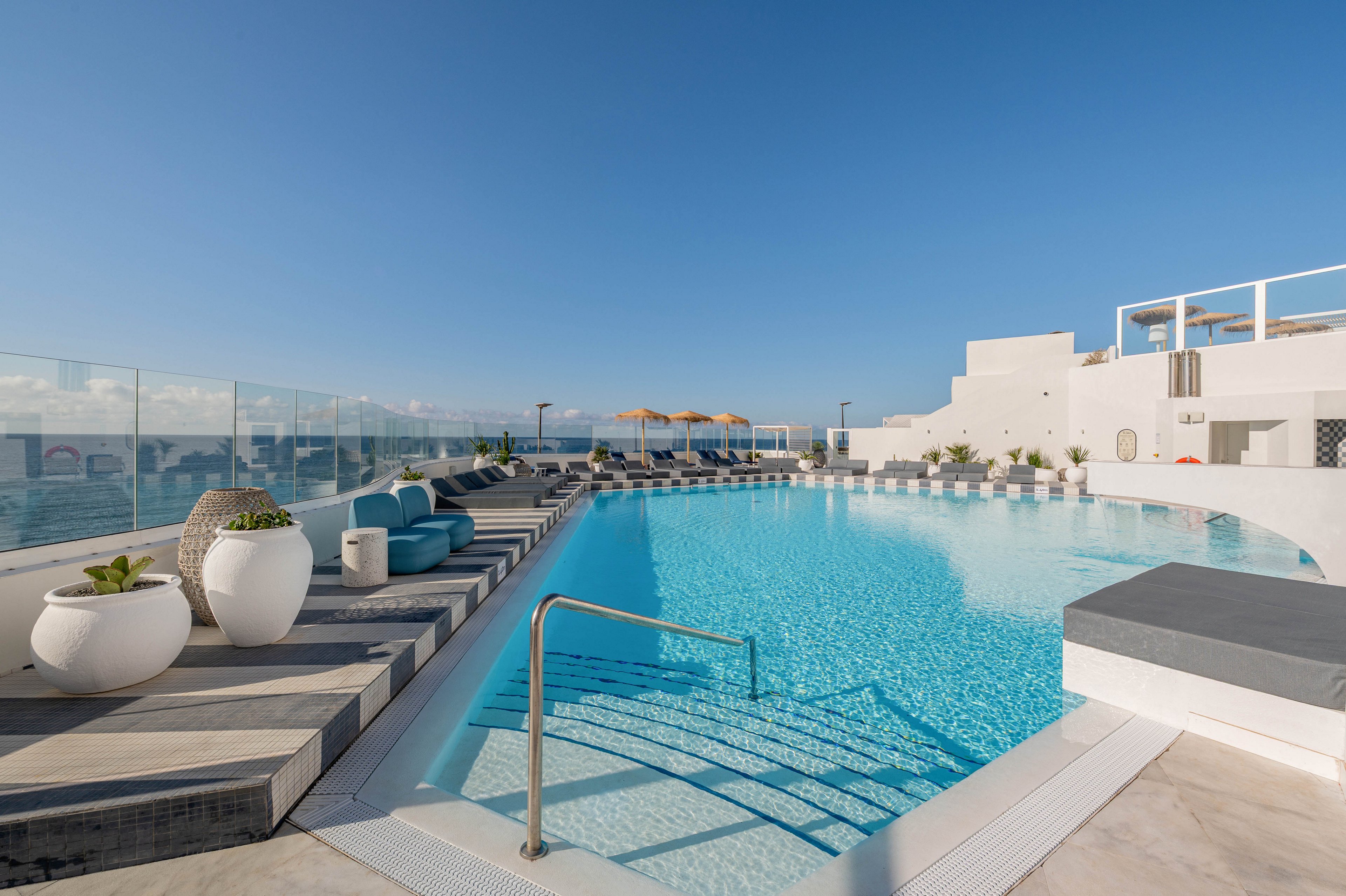 A contemporary rooftop infinity pool stretches alongside a glass railing with views of the ocean. Sun loungers and umbrellas line the far side of the pool, creating a relaxing resort atmosphere. Large white planters with green plants decorate the deck, and clean architectural lines emphasize the modern design. The clear blue water and bright sky contribute to a calm, luxurious vacation setting.
