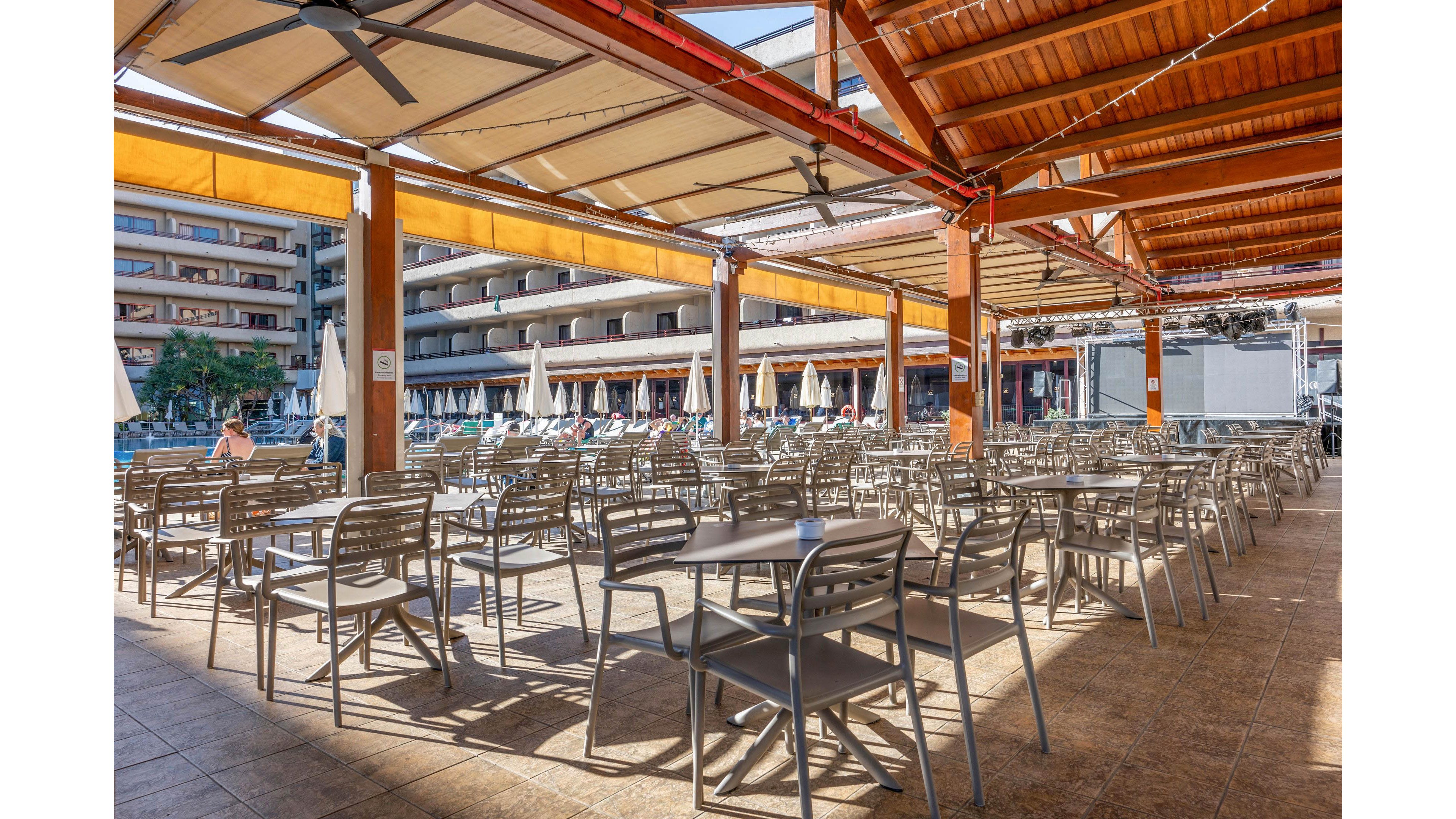 The image shows a spacious outdoor terrace filled with rows of empty tables and chairs. A wooden pergola with retractable awnings provides shade over the seating area. In the background, a hotel or apartment building with balconies overlooks a swimming pool lined with sun loungers and umbrellas. Bright daylight and clear skies create a relaxed vacation atmosphere.