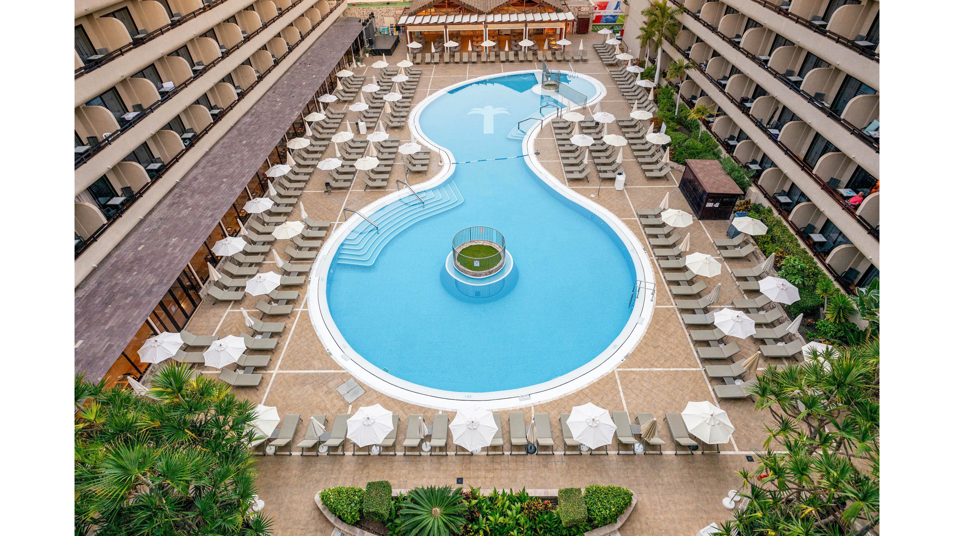 An aerial view shows a large, uniquely shaped swimming pool centered in a hotel courtyard. Rows of sun loungers and white umbrellas line both sides of the pool, creating a symmetrical, organized layout. Multi-story balconies overlook the pool area, while lush green plants and palm trees add a tropical touch around the edges. The scene suggests a resort-style setting designed for relaxation and leisure.