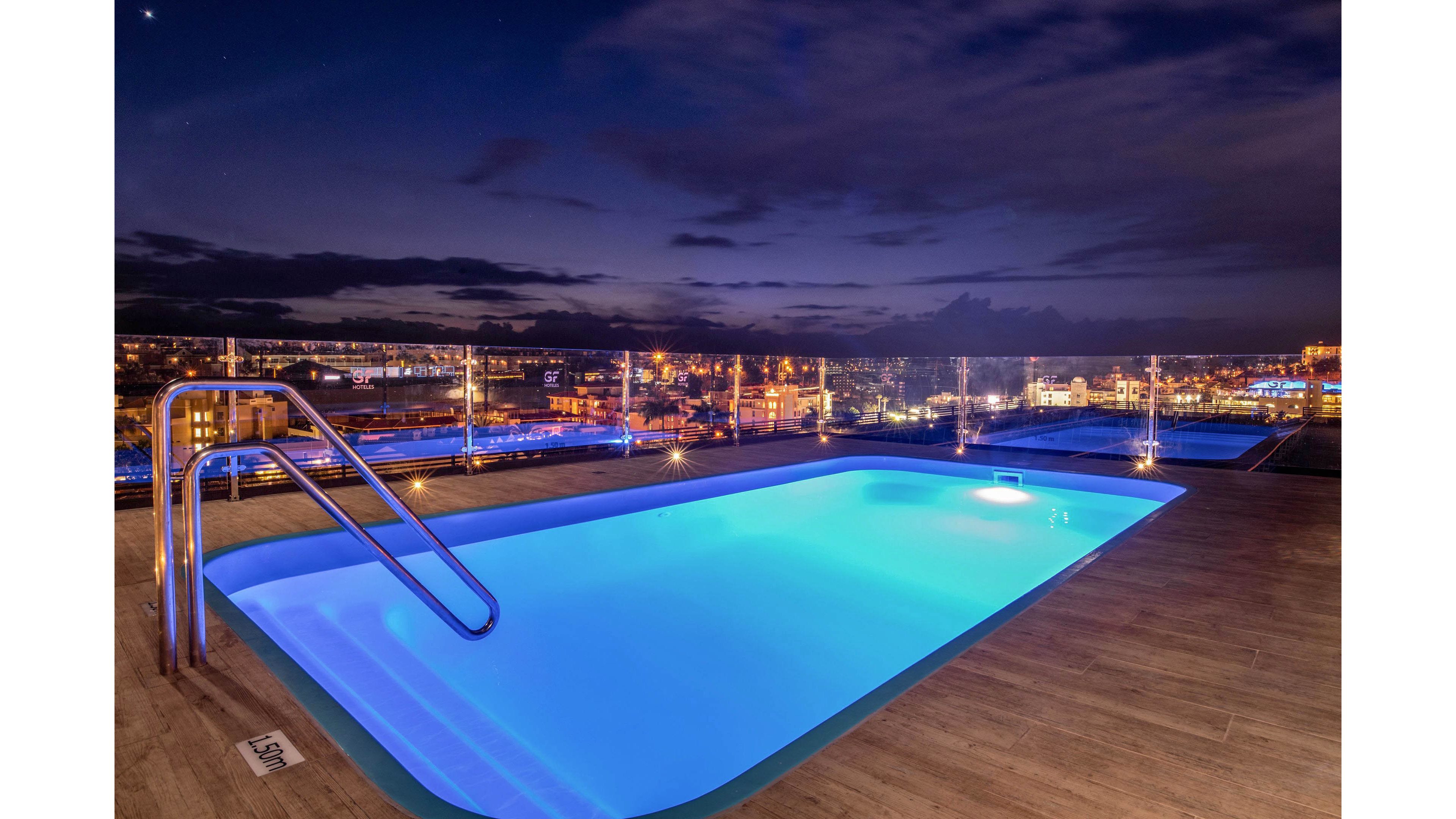 A rooftop swimming pool glows with bright blue lighting against a dark night sky. Stainless steel handrails lead into the water, surrounded by a wooden deck. Glass railings frame the terrace, revealing a cityscape illuminated with warm lights in the distance. The calm atmosphere and reflections create a luxurious, tranquil nighttime setting.