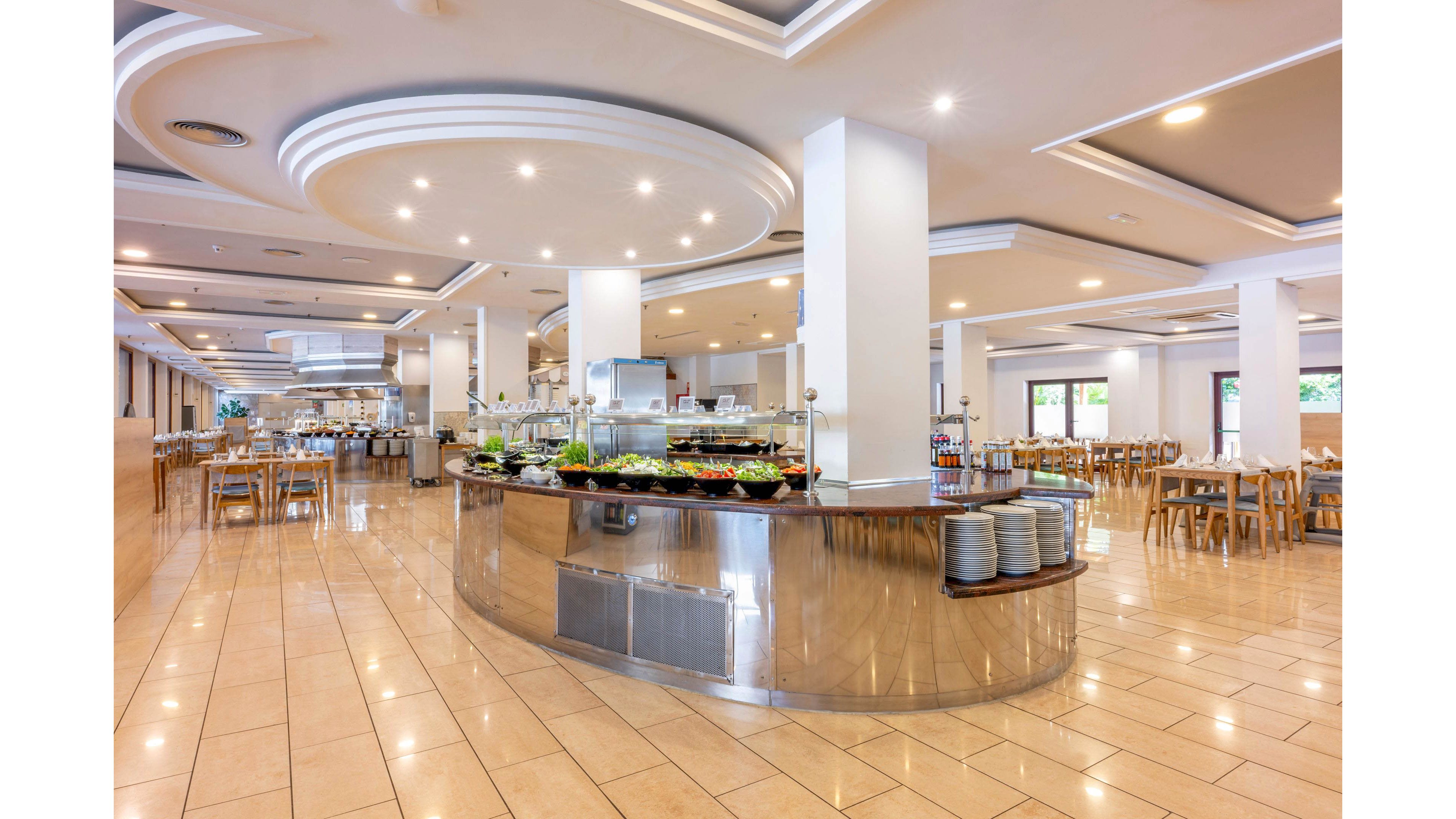 A spacious self-service buffet area stands in the center of a modern restaurant. The room features glossy tiled floors, white columns, and recessed ceiling lights that create a bright, clean atmosphere. Tables and wooden chairs are neatly arranged around the buffet, ready for guests. Fresh food trays and stacked plates on the counter emphasize a prepared dining service setting.