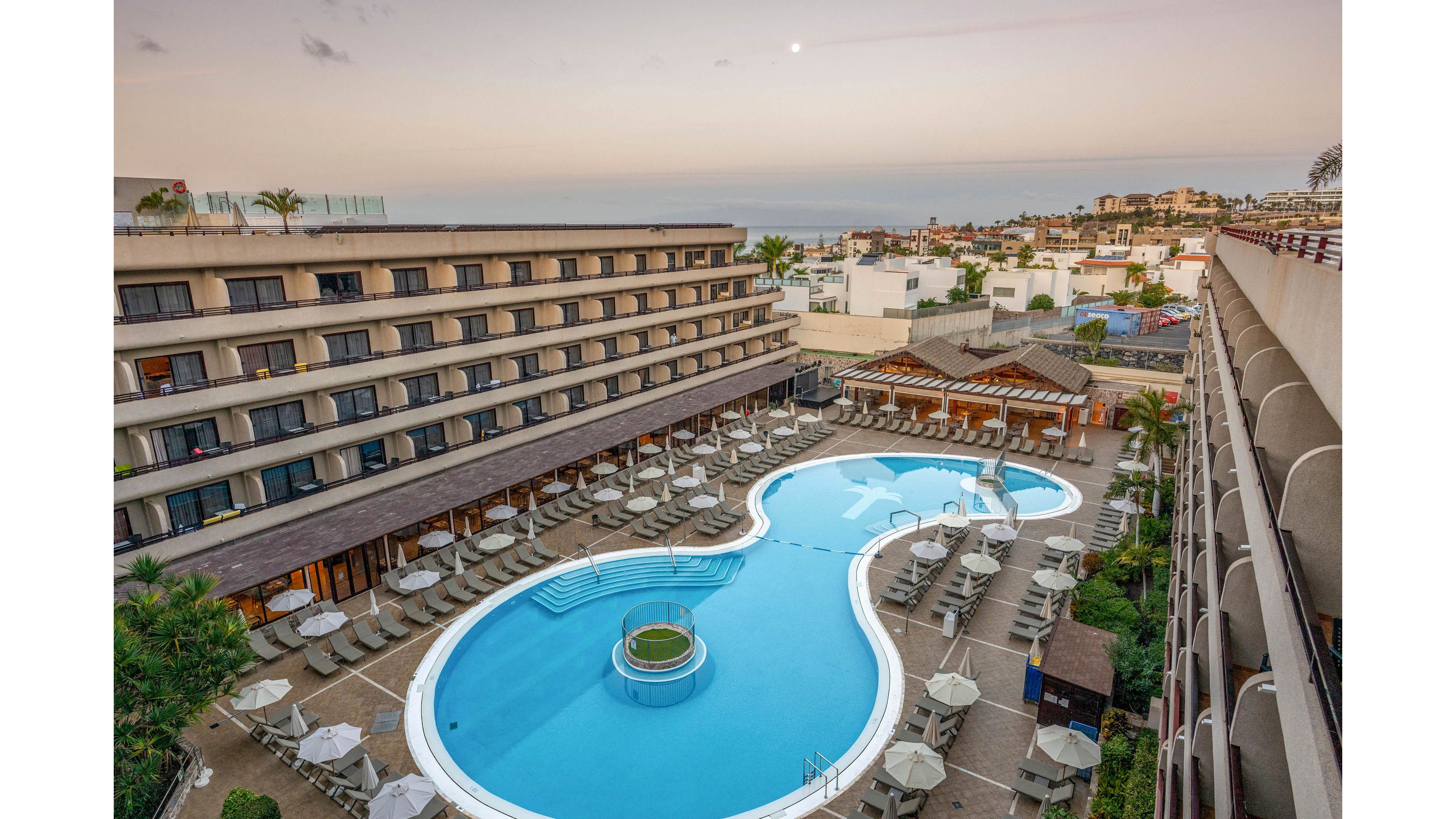 An elevated view shows a large resort hotel with a uniquely shaped outdoor swimming pool at its center. Numerous sun loungers and umbrellas surround the pool, arranged in neat rows on the terrace. The multi-story hotel buildings frame the pool area, with balconies overlooking the scene. In the distance, a coastal town and the sea are visible under a soft evening or early morning sky.