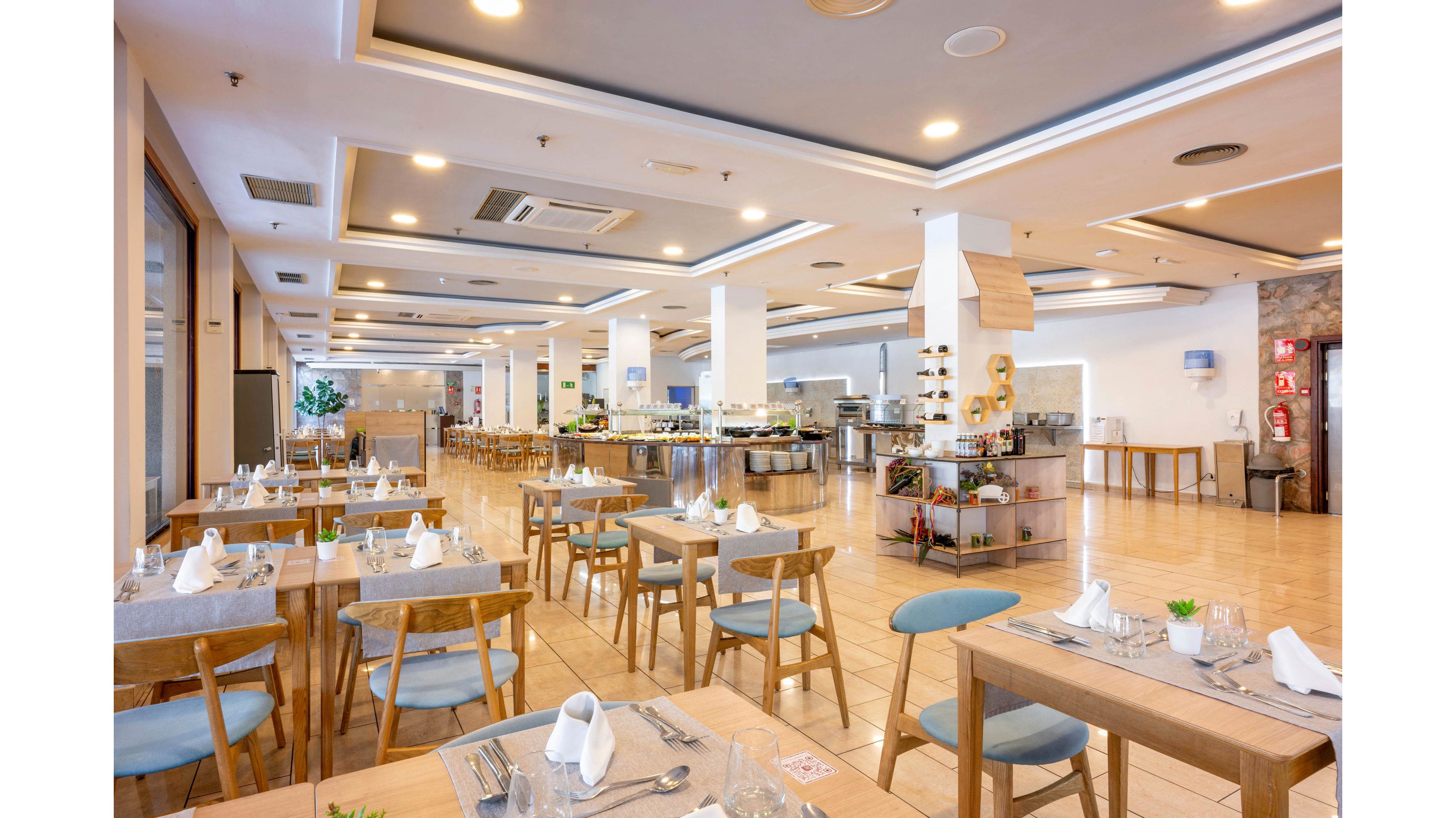 Spacious modern restaurant interior with neatly arranged wooden tables and chairs. Tables are set with white napkins, glassware, and cutlery, ready for guests. A large buffet area with counters and food displays extends through the center of the room. Warm lighting, light wood flooring, and neutral tones create an inviting, contemporary atmosphere.
