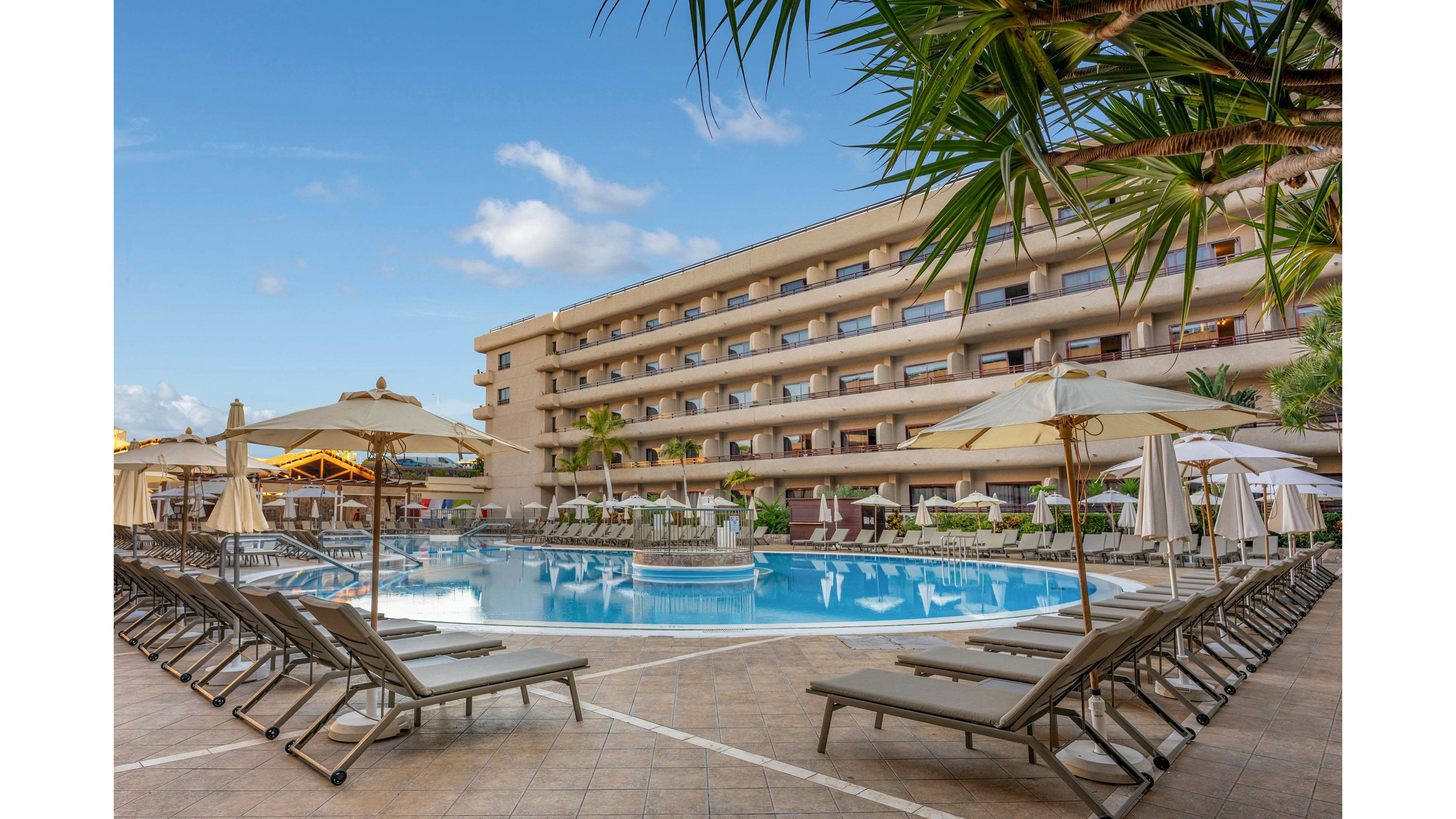 A large resort hotel features a central outdoor swimming pool surrounded by multiple stories of balconies. Numerous sun loungers and umbrellas line the tiled pool deck, creating a relaxing vacation atmosphere. Palm trees and landscaped greenery frame the scene under a clear blue sky with a few clouds. Warm lighting from the building and pool area suggests an inviting, tranquil evening setting.