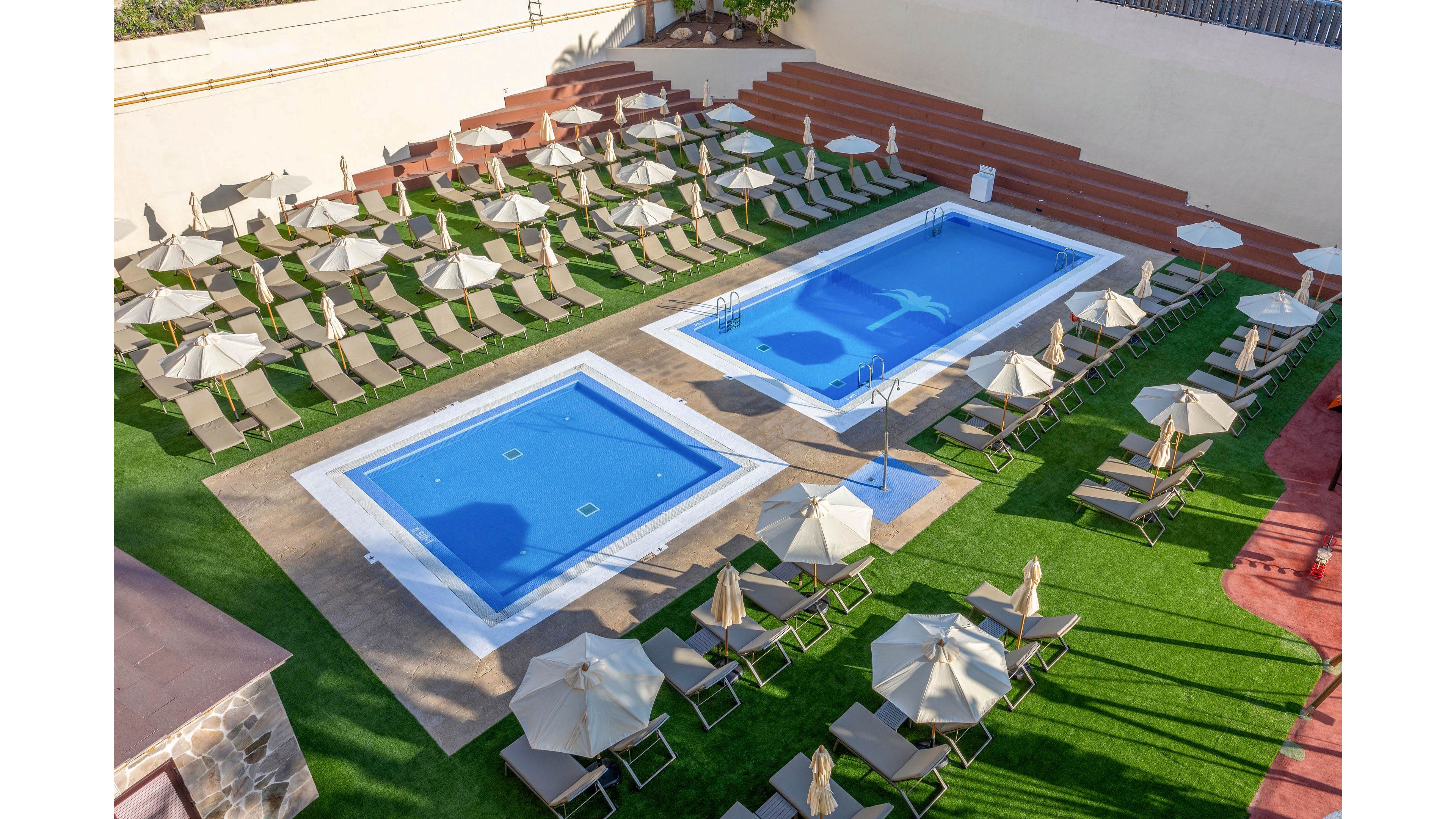A pair of rectangular blue swimming pools sit in the center of a resort sun deck. Numerous beige sun loungers and umbrellas are neatly arranged around the pools on artificial grass. The area is empty of people, suggesting early morning or off-peak hours. Surrounding walls and tiered seating frame the orderly, relaxing outdoor space.