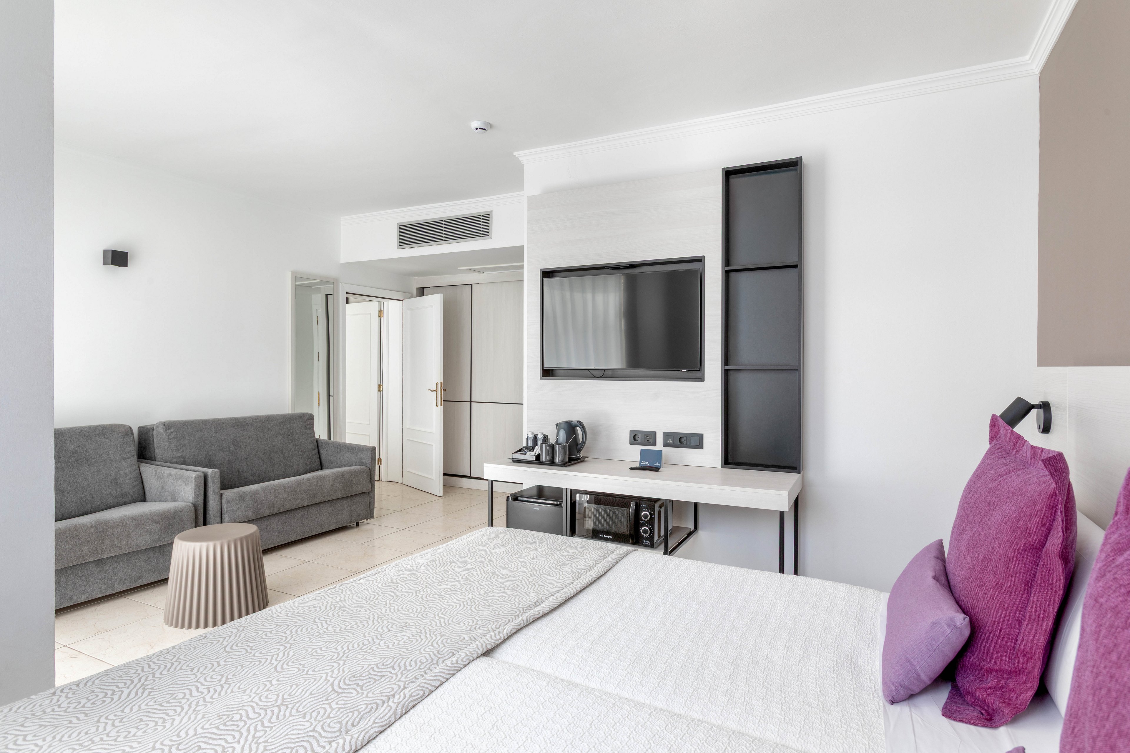 A contemporary hotel room features a neatly made bed with light bedding and purple accent pillows in the foreground. The space includes a gray sofa and a small ribbed side table, creating a comfortable seating area. A wall-mounted flat-screen television is positioned above a desk with a kettle and cups, suggesting in-room refreshments. White walls, tiled flooring, and minimal decor give the room a clean, airy, and modern feel.