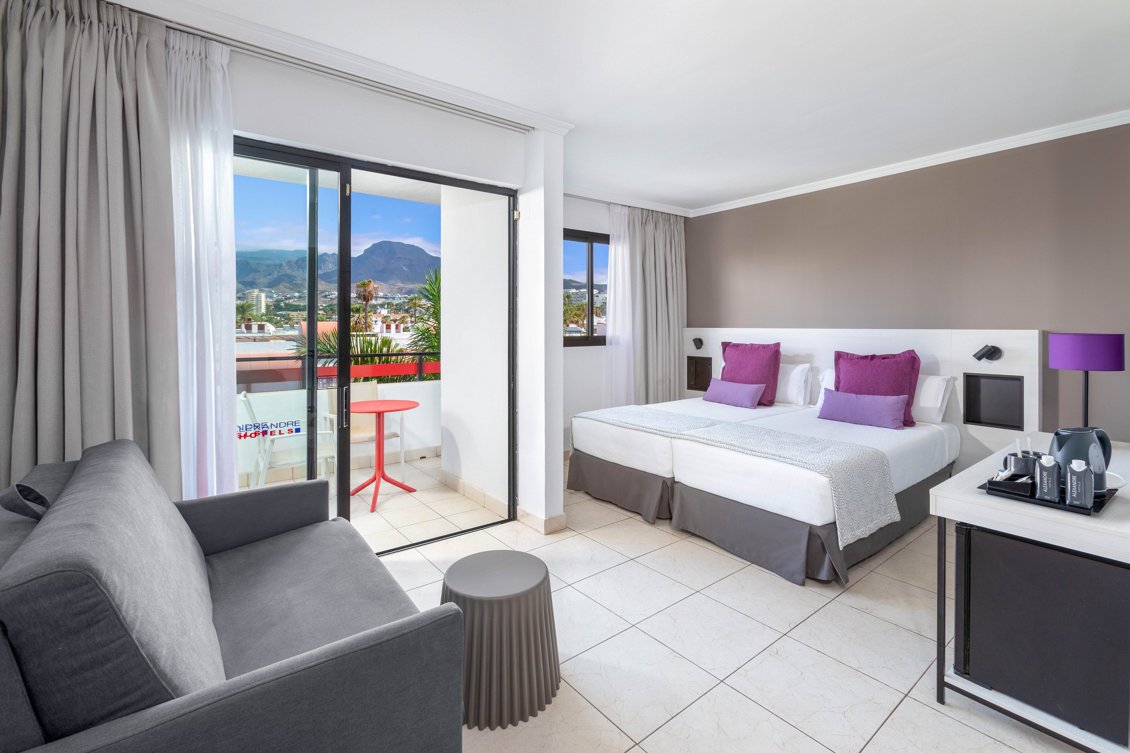 Bright contemporary hotel room featuring two single beds with white linens and purple cushions. A gray sofa and small round side table sit opposite a compact refreshment station with a coffee machine. Large sliding glass doors open to a balcony with a red table and chairs, overlooking a town and distant mountains. Neutral walls and tiled floors create a clean, minimalist atmosphere with purple accent decor.