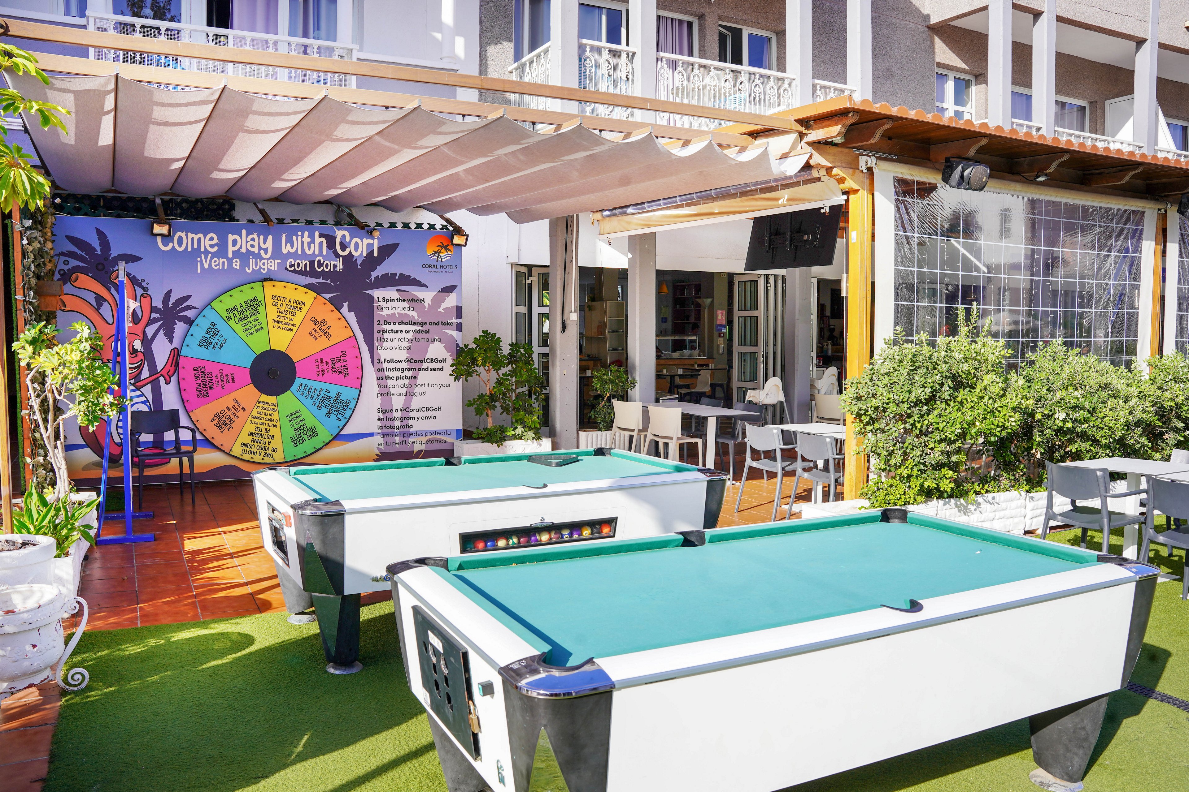 An outdoor leisure area features two white pool tables set on artificial grass in front of a hotel or resort building. Behind the tables, a colorful game wheel display reads "Come play with Cort" and lists various playful activities. The space is shaded by a pergola canopy and surrounded by potted plants and greenery, with chairs and tables for guests. Bright daylight and vivid colors create a fun, family-friendly atmosphere.