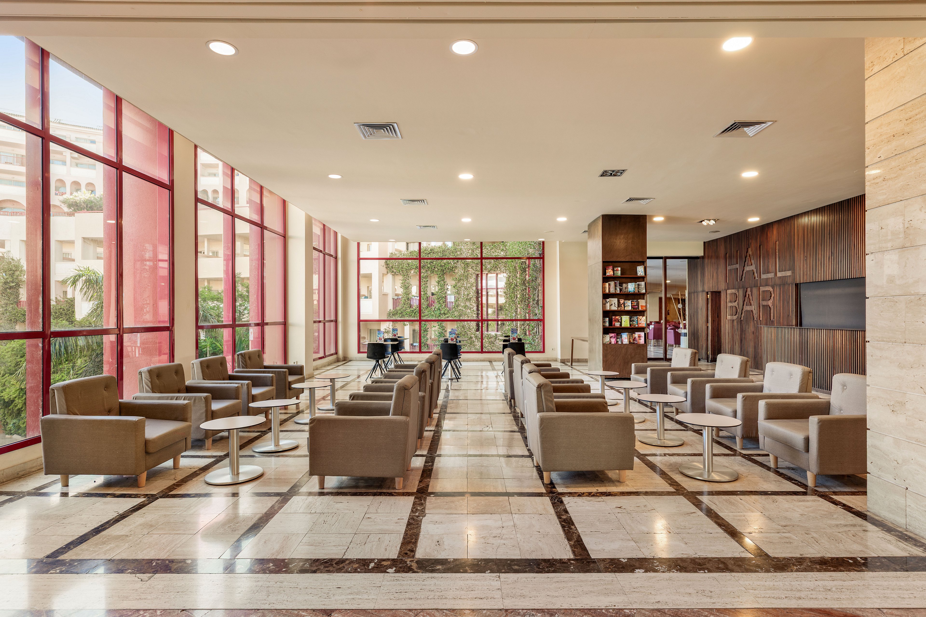 A spacious hotel lobby lounge features rows of modern armchairs and small round tables arranged symmetrically. Large floor-to-ceiling windows with red frames allow natural light to flood the space and reveal greenery outside. The polished marble floor and recessed ceiling lights create a bright, contemporary atmosphere. At the far end, a bar area and shelving unit add a social focal point to the otherwise quiet seating area.