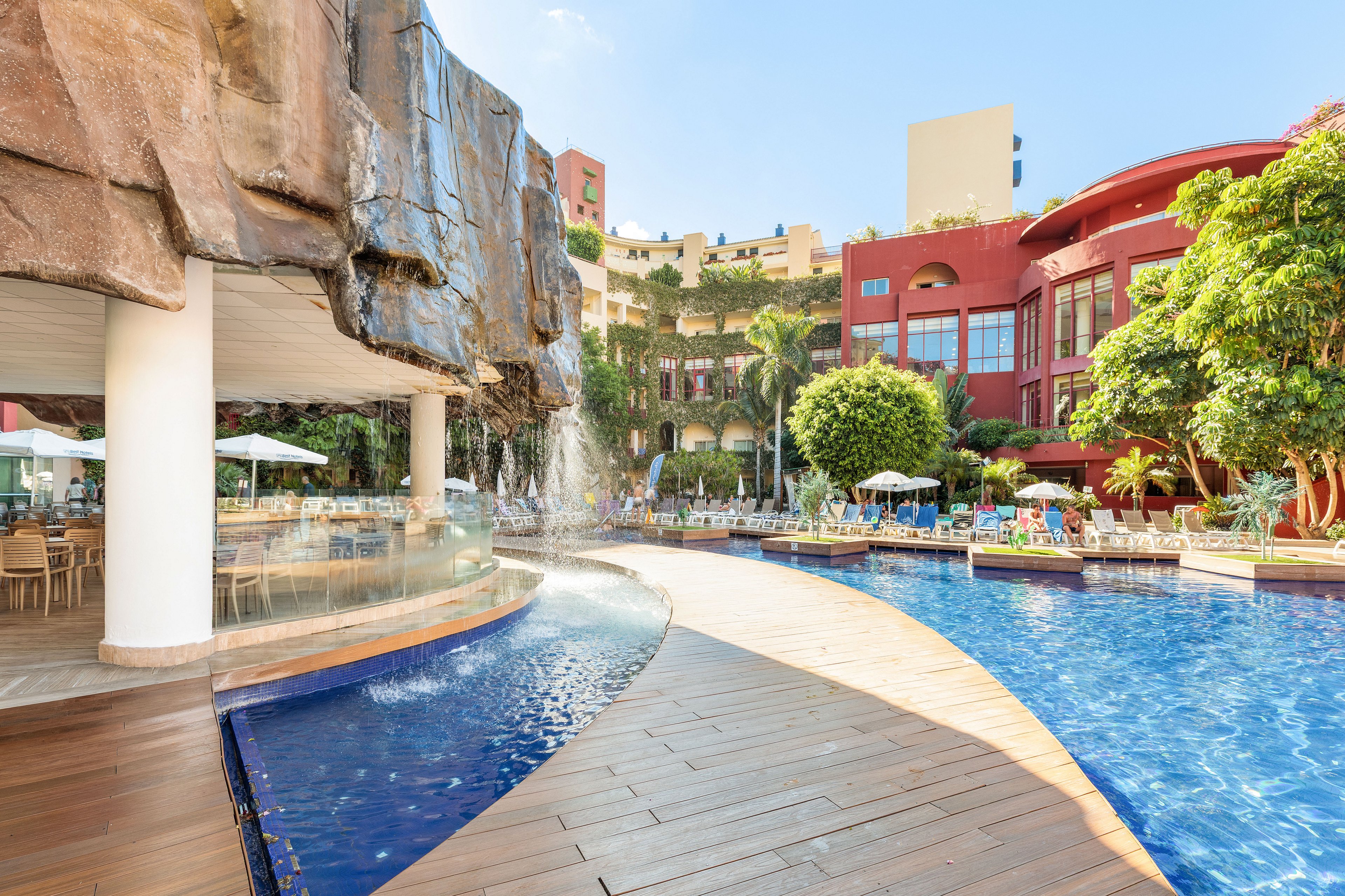 A large resort swimming pool curves around a rock formation with a cascading waterfall. The scene includes a wooden deck walkway that follows the edge of the pool. In the background, guests relax on sun loungers near palm trees and lush greenery. Colorful multi-story buildings frame the pool area under a clear blue sky, creating a bright and inviting vacation atmosphere.