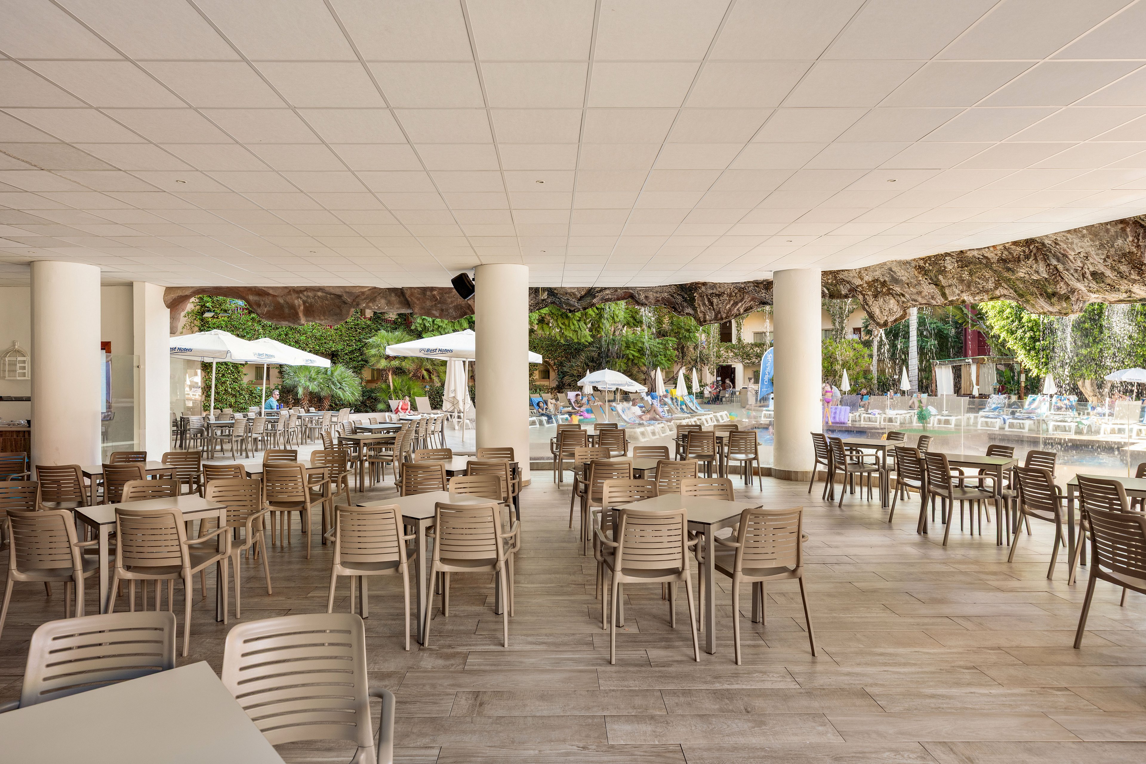 An open-air terrace café features rows of empty tables and chairs under a white ceiling. The space overlooks a resort swimming pool area with sun loungers and large umbrellas. Natural rock formations and lush greenery frame the background, creating a relaxed vacation atmosphere. Bright daylight and neutral tones give the scene a clean, inviting look.