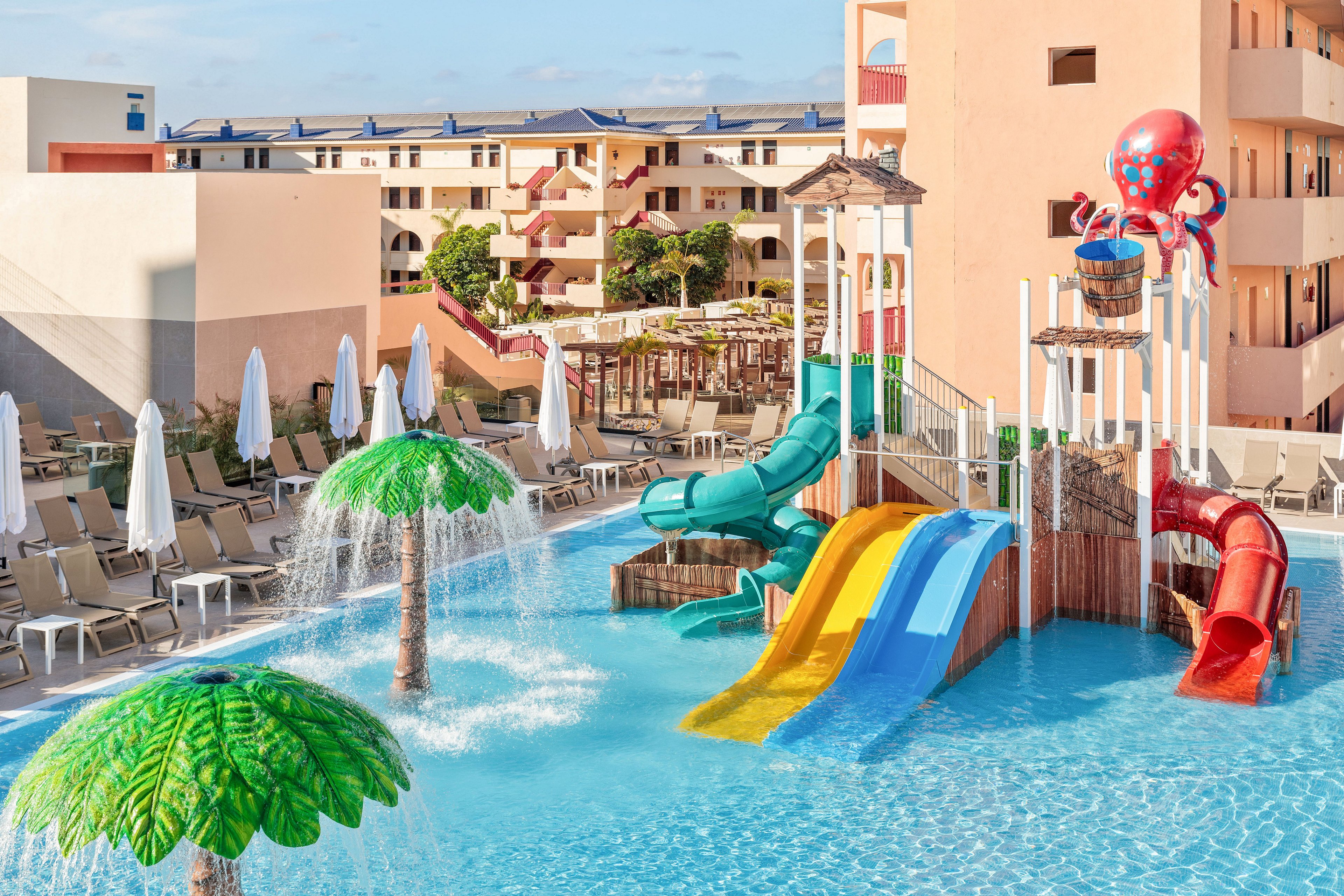A vibrant children’s water playground features multiple colorful slides leading into a shallow pool. Palm tree fountains and mushroom-shaped sprayers create playful streams of water. An octopus-themed structure with a tipping bucket adds to the fun, set against a backdrop of resort buildings and sun loungers. The scene is bright and sunny, suggesting a family-friendly vacation destination.