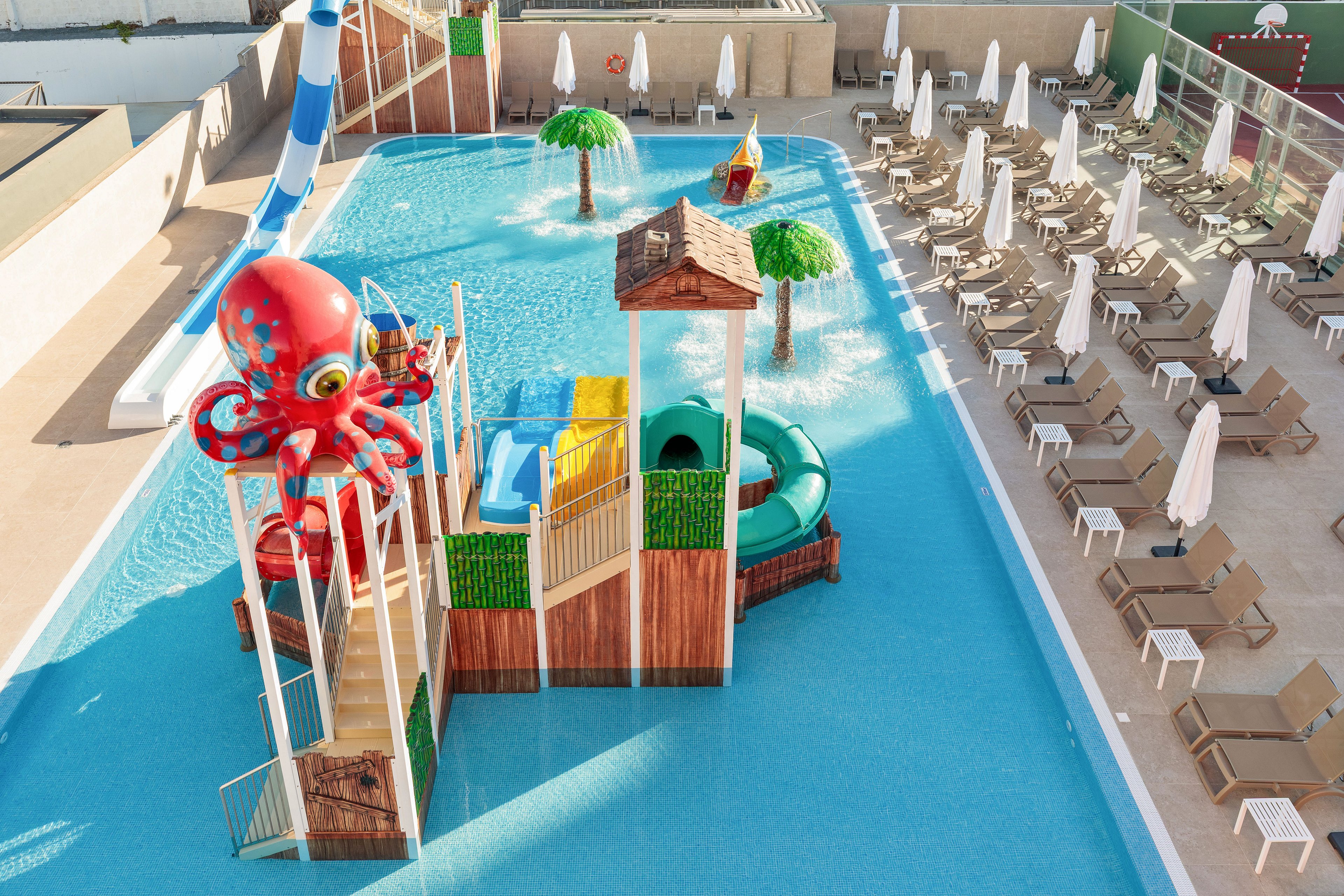 A bright children’s water playground features a large red octopus structure in a shallow resort pool. Slides, climbing platforms, and small play towers are surrounded by clear blue water. In the background, rows of sun loungers with white umbrellas line the pool deck. The scene suggests a family-friendly water park area designed for kids’ recreation and relaxation.