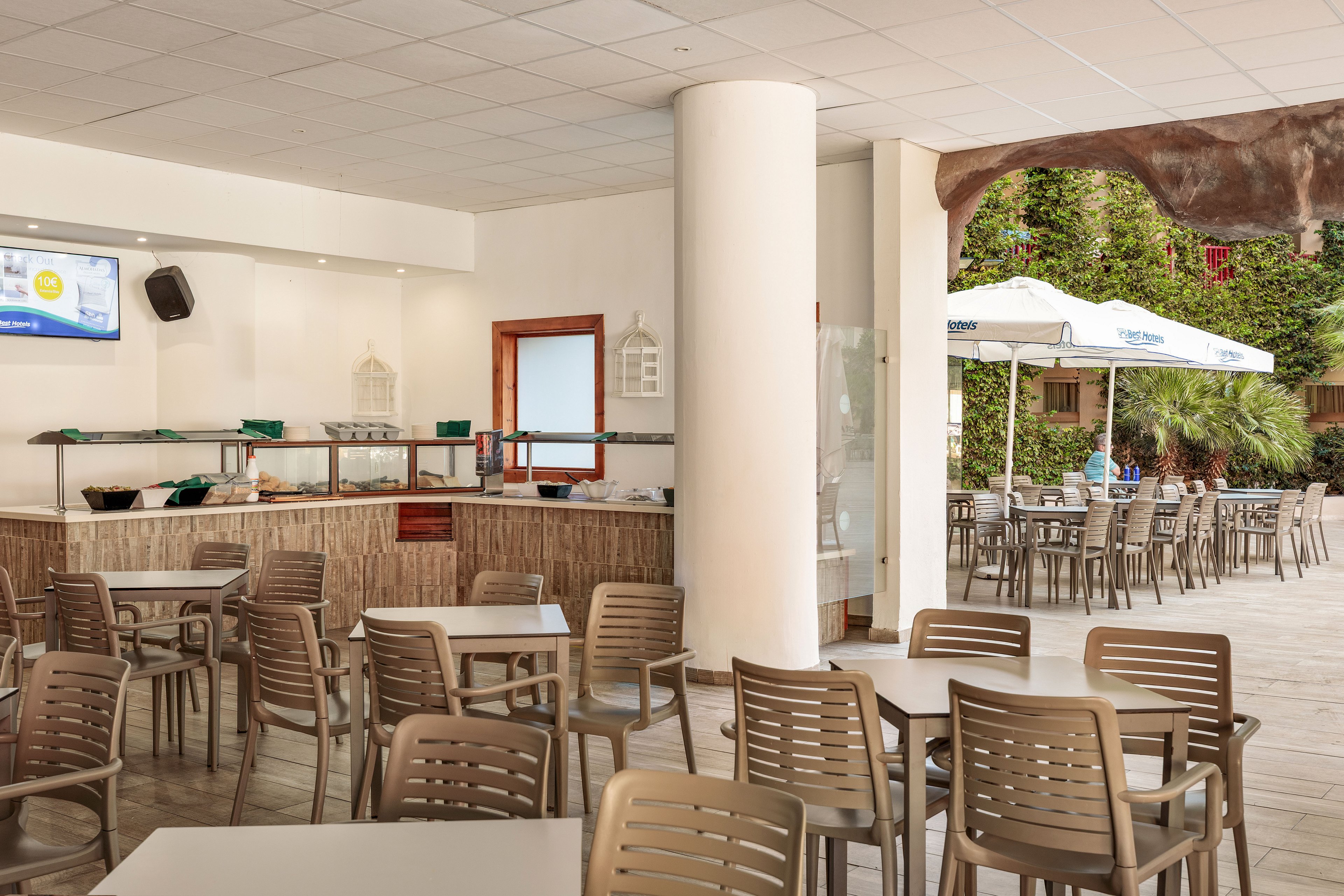 An open-air cafe area features rows of empty plastic tables and chairs arranged around a central column. A buffet-style counter with glass sneeze guards and a mounted television screen stands along the back wall. Large umbrellas shade additional seating on the patio outside, surrounded by greenery and palm trees. The bright, neutral color palette and clean layout suggest a relaxed, casual dining environment.