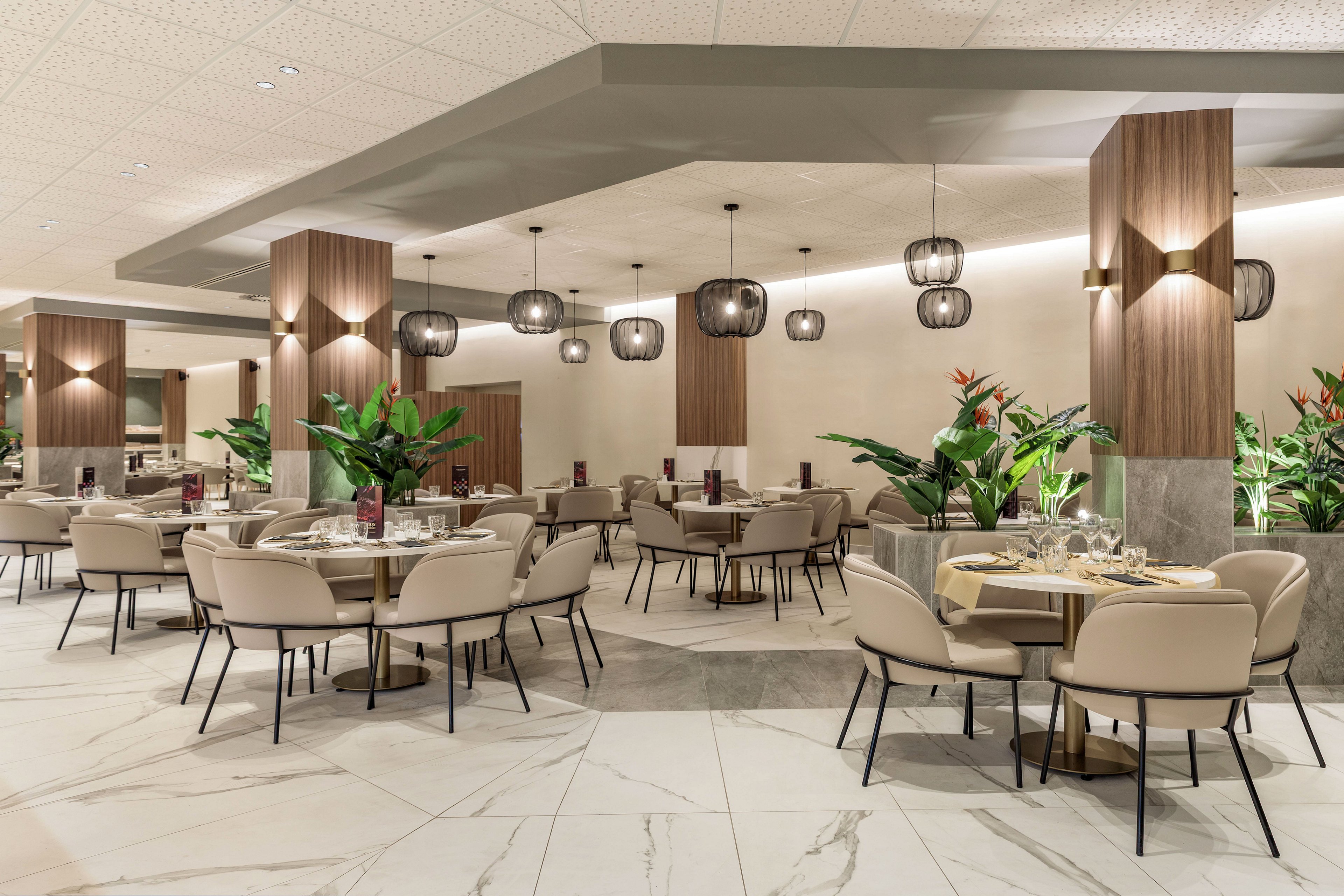 A contemporary restaurant dining room features round tables set with plates, glasses, and folded napkins. Soft beige chairs surround the tables, arranged neatly across a spacious marble-tiled floor. Warm wood paneling, large potted plants, and sculptural pendant lights create a sophisticated atmosphere. The space appears calm and inviting, designed for comfortable, upscale dining.