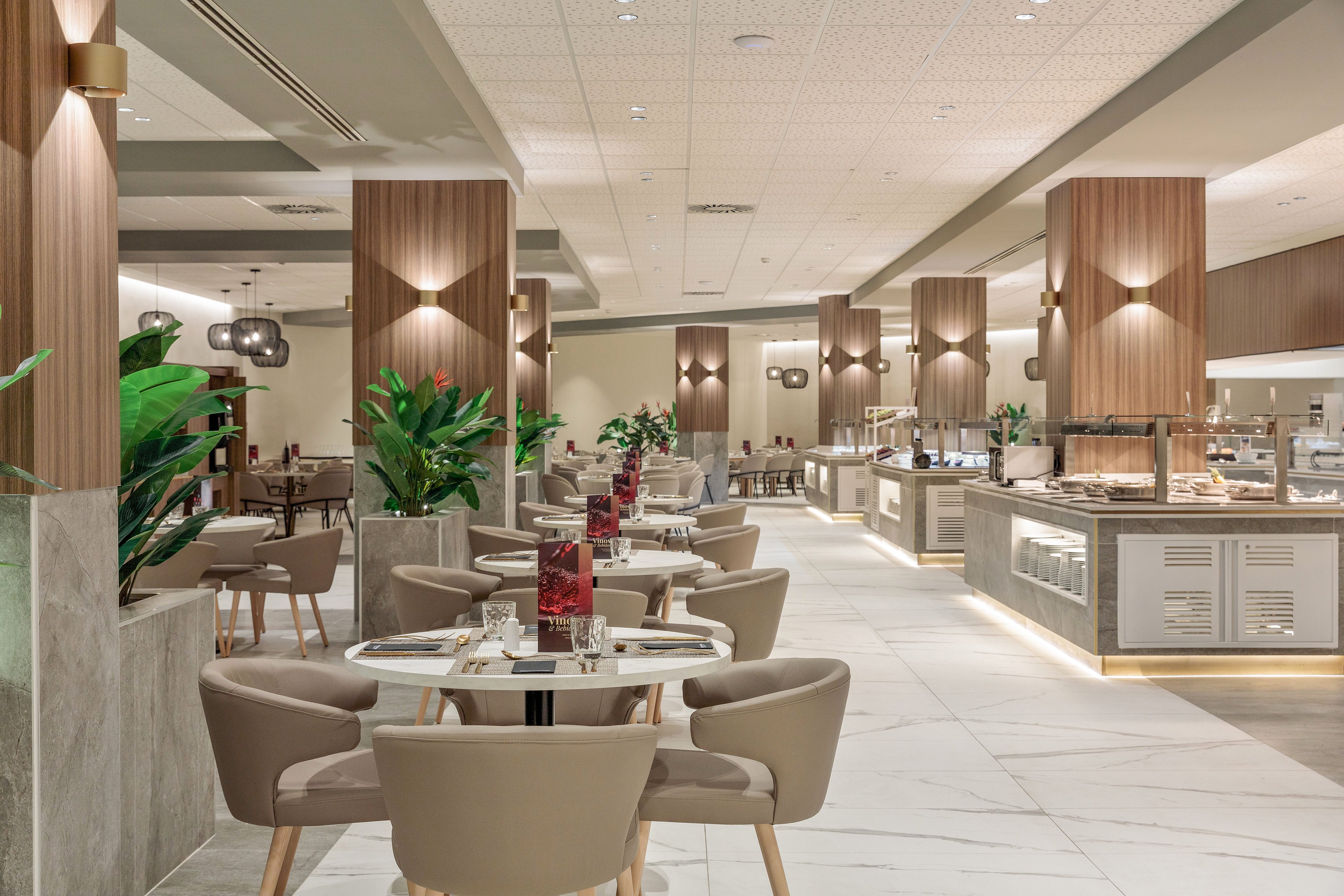 A contemporary restaurant interior features neatly arranged round tables with beige upholstered chairs. The setting appears to be a hotel buffet area, with multiple food stations and counters visible in the background. Warm wooden columns, soft lighting, and large green plants create a welcoming and sophisticated atmosphere. The space is clean, spacious, and designed with neutral tones and minimalist decor.