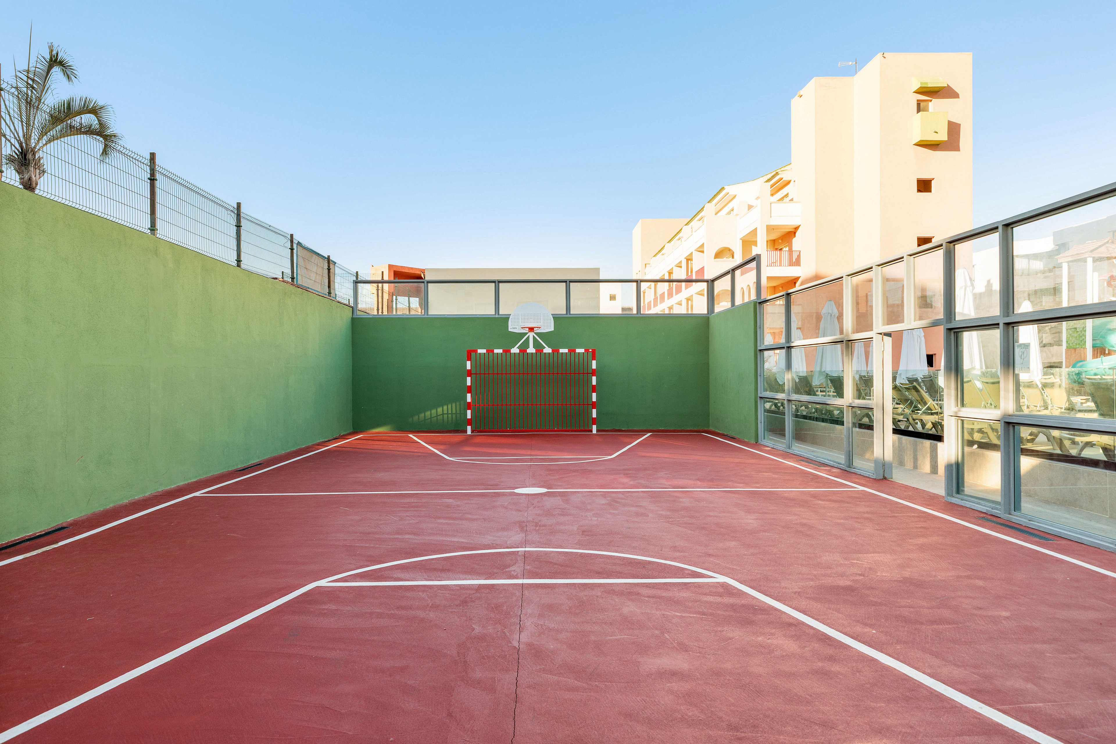 An empty outdoor sports court with red flooring and white lines is shown under a clear blue sky. The court is enclosed by green and glass walls, with a small goal and basketball hoop at one end. Residential or hotel buildings are visible in the background, suggesting an urban or resort setting. The warm sunlight creates long shadows and a calm, peaceful atmosphere.