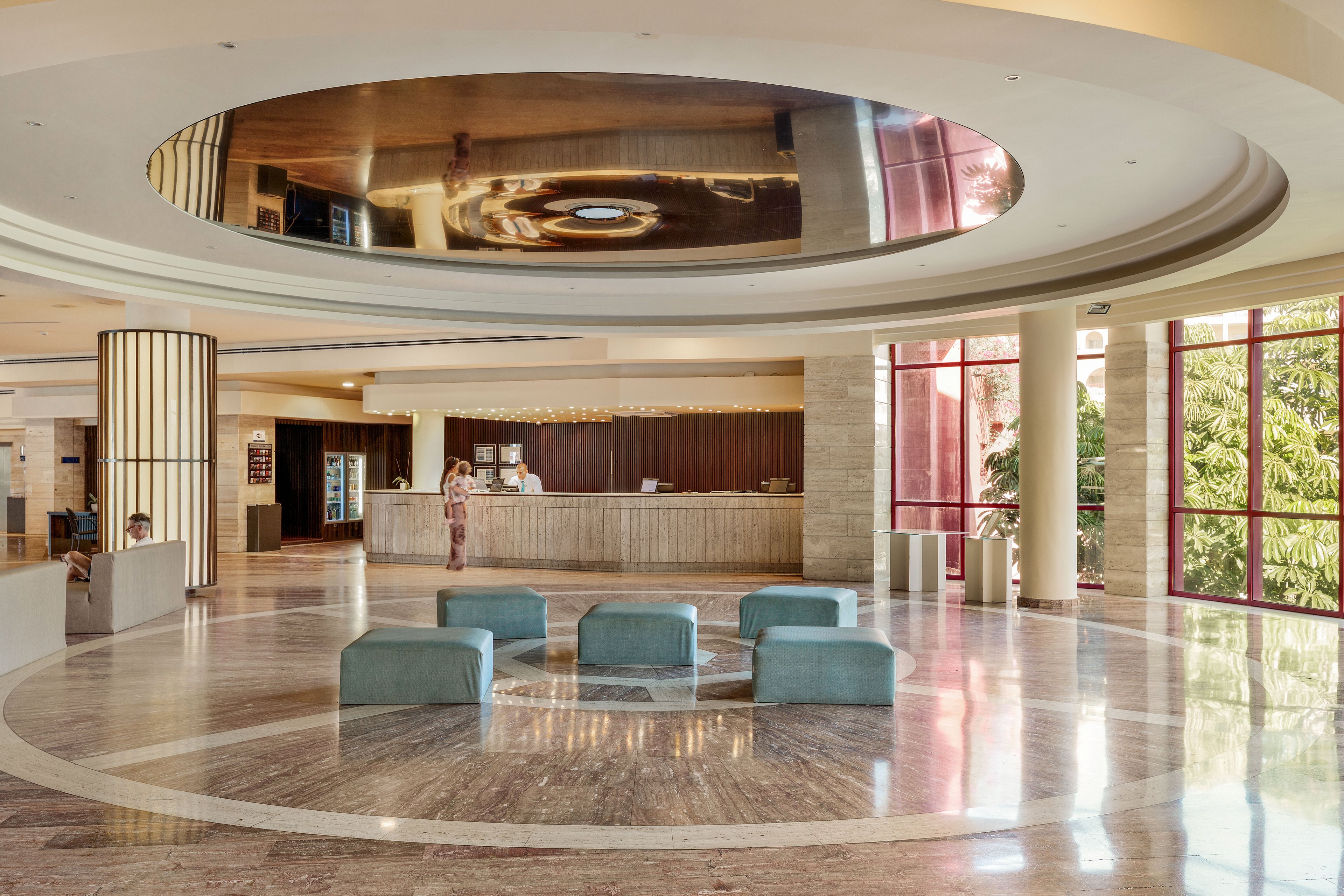 A spacious, contemporary hotel lobby features a circular marble floor and a reflective ceiling detail. Several teal upholstered ottomans are arranged in the center of the open space. In the background, a guest stands at the reception desk while staff assist from behind the counter. Large floor-to-ceiling windows on the right bring in natural light and views of greenery, enhancing the airy, welcoming atmosphere.