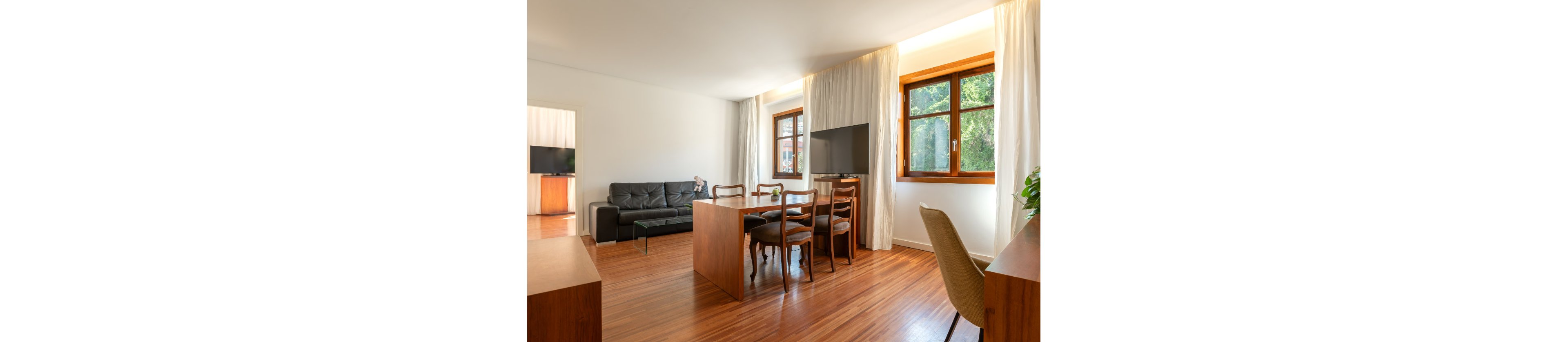A bright modern living room opens into a dining area with wooden furniture and a sleek black sofa. Large windows with white curtains let in natural light and reveal greenery outside. A flat-screen television is mounted on a stand near the dining table, while a desk and chair sit along the opposite wall. The space features warm wooden flooring, minimal decor, and a clean contemporary style.