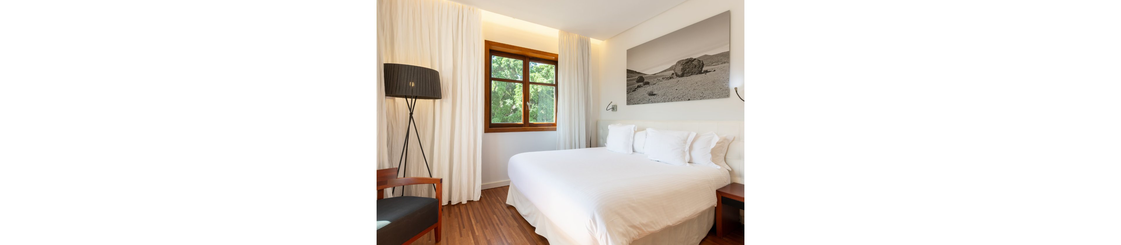A bright, modern bedroom features a large neatly made bed with white linens and multiple pillows. Natural light streams through a wooden-framed window, revealing green trees outside. White curtains, a black floor lamp, and a wooden chair add contrast to the minimalist decor. A large black-and-white landscape photograph hangs above the bed, enhancing the serene and contemporary atmosphere.