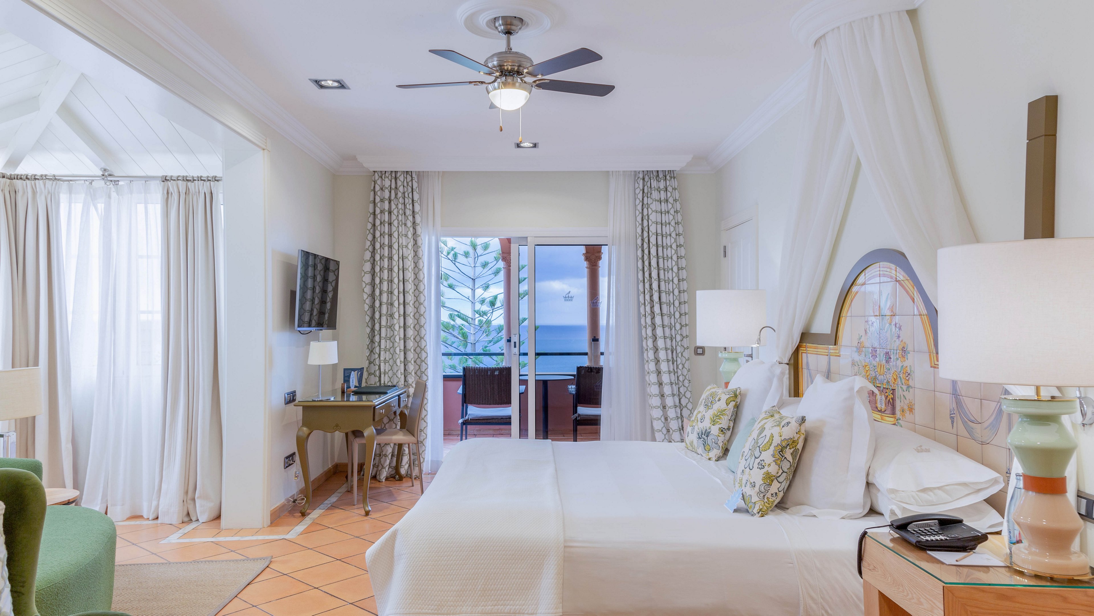 A spacious hotel bedroom features a large neatly made bed with white linens and patterned pillows. The room is decorated in soft neutral tones with warm tile flooring, bedside lamps, and a ceiling fan above. A desk with a television and chair sits along one wall, while sheer curtains frame a doorway leading to a balcony. Through the open doors, an outdoor seating area and a view of the ocean and trees are visible, creating a serene coastal atmosphere.