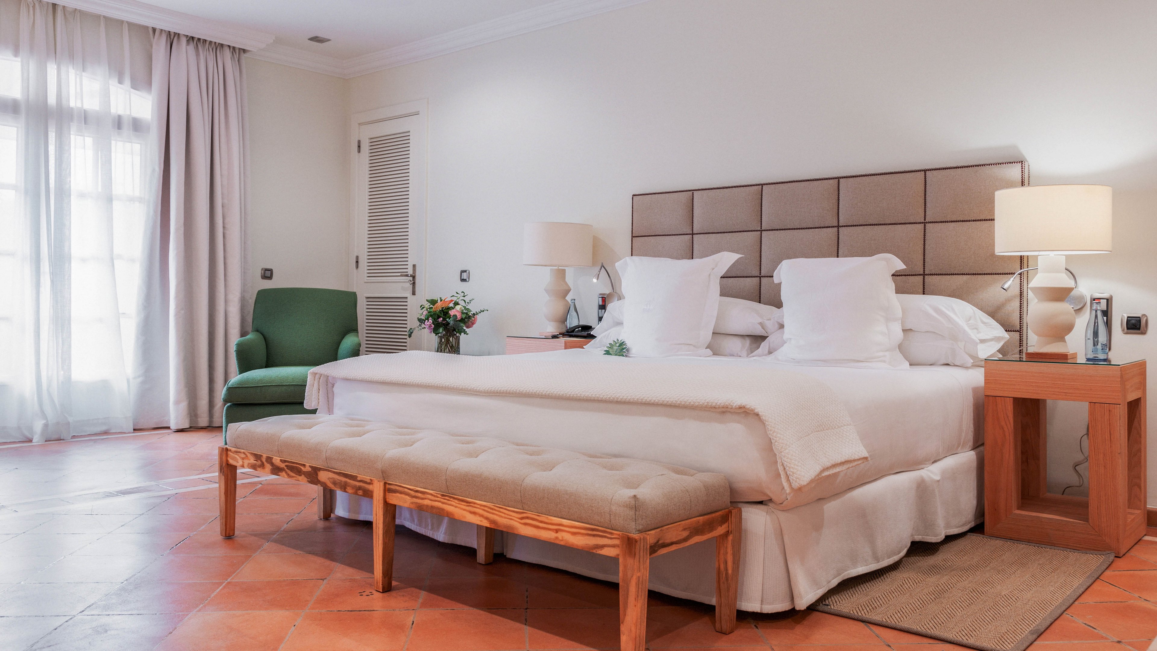 A spacious, contemporary bedroom features a large neatly made bed with multiple white pillows and a padded headboard. A cushioned bench sits at the foot of the bed, while matching wooden nightstands with table lamps flank each side. A green upholstered armchair and side table with flowers create a cozy seating corner near a window with sheer curtains. Warm terracotta floor tiles and soft neutral walls contribute to a calm, inviting atmosphere.