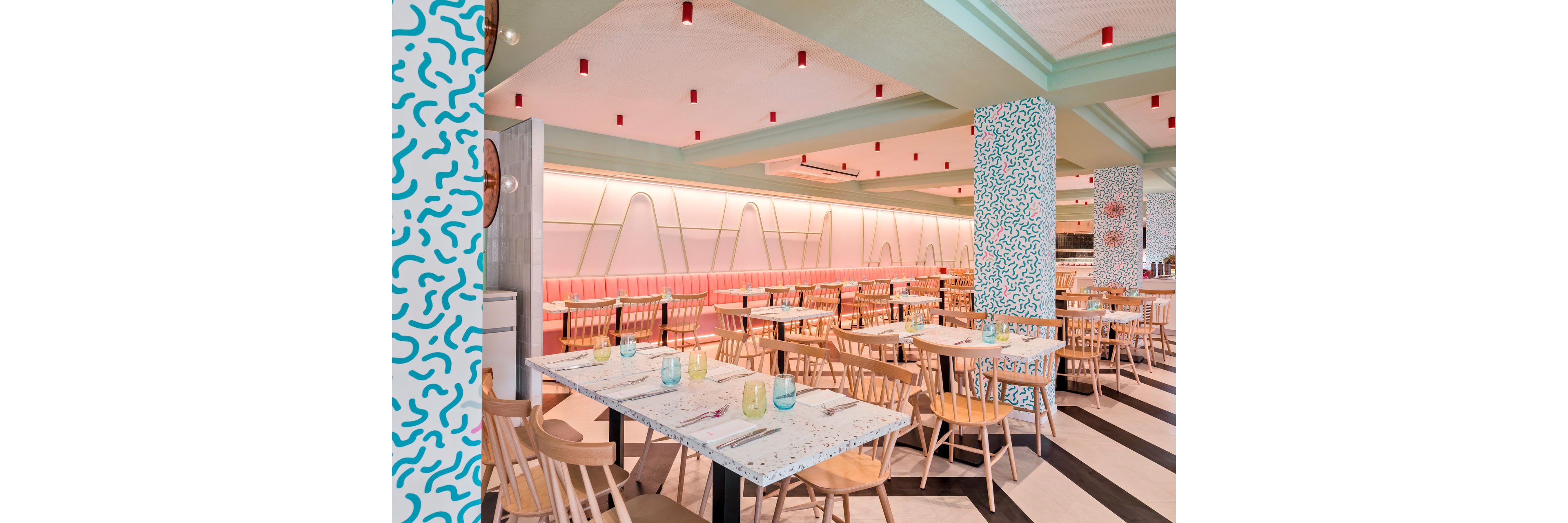 Bright contemporary restaurant interior featuring rows of wooden chairs and light-colored tables. The space is decorated with pastel pink banquettes, mint green accents, and playful patterned columns. Striped flooring and red ceiling lights add graphic contrast to the design. Glasses and cutlery are neatly arranged on the tables, suggesting a ready-to-serve dining area.