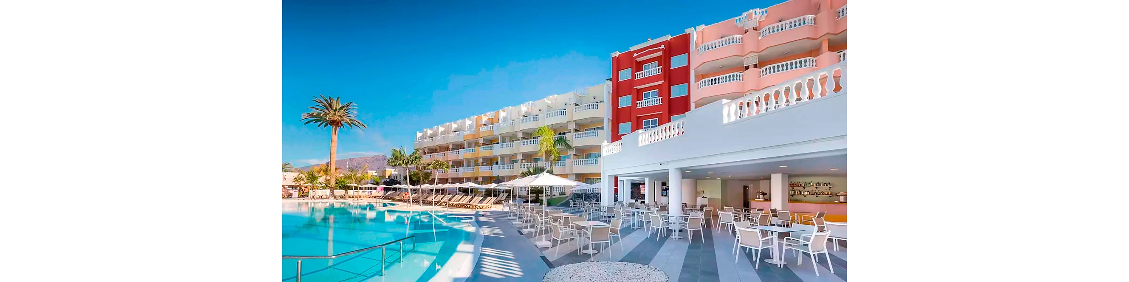 A large outdoor swimming pool sits beside a bright multi-story resort complex with colorful facades. White lounge chairs and umbrellas line the pool deck, leading to a spacious terrace filled with tables and chairs. A bar area is visible under a covered section of the building, suggesting a relaxed vacation setting. Palm trees and clear blue sky enhance the tropical, sunny atmosphere.