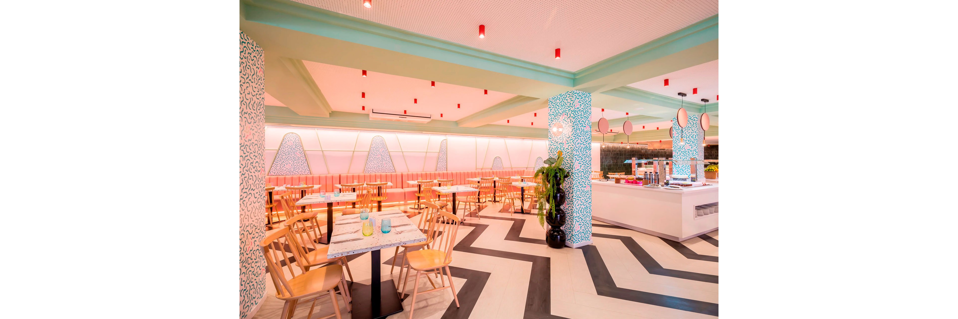 Contemporary restaurant dining area featuring light wooden chairs and simple tables is shown in a brightly lit space. The setting includes pastel pink and mint green walls, patterned columns, and geometric black-and-white floor tiles. Red ceiling lights and decorative wall panels add playful accents to the clean, modern design. A counter area and indoor plants complete the fresh, inviting atmosphere.
