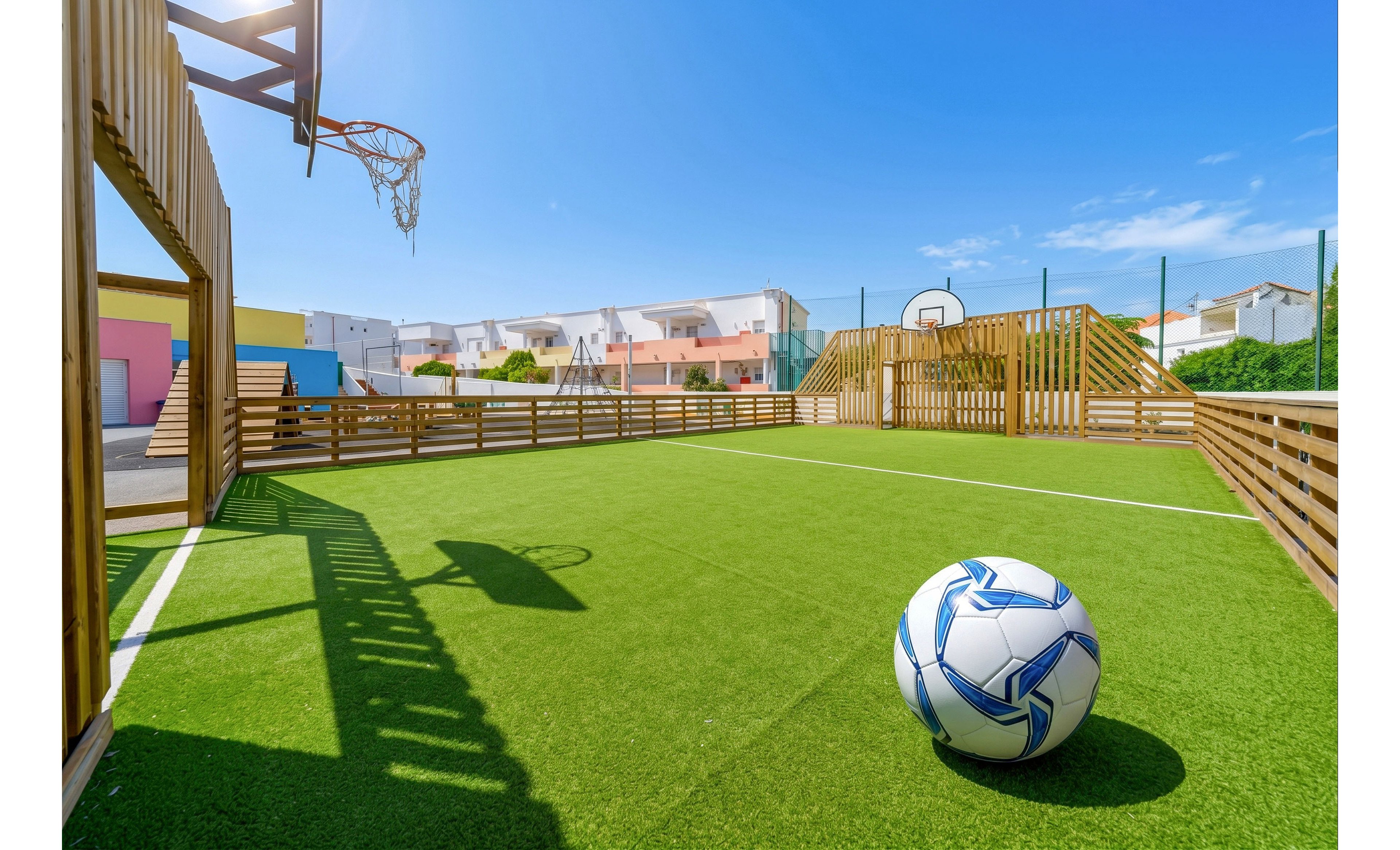 An outdoor sports court with bright green artificial turf features a white and blue soccer ball in the foreground. Wooden fences and structures surround the playing area, with a basketball hoop visible in the background. Residential buildings and a clear blue sky frame the scene, suggesting a neighborhood recreational space. Strong sunlight creates sharp shadows, giving the image a vibrant and energetic feel.