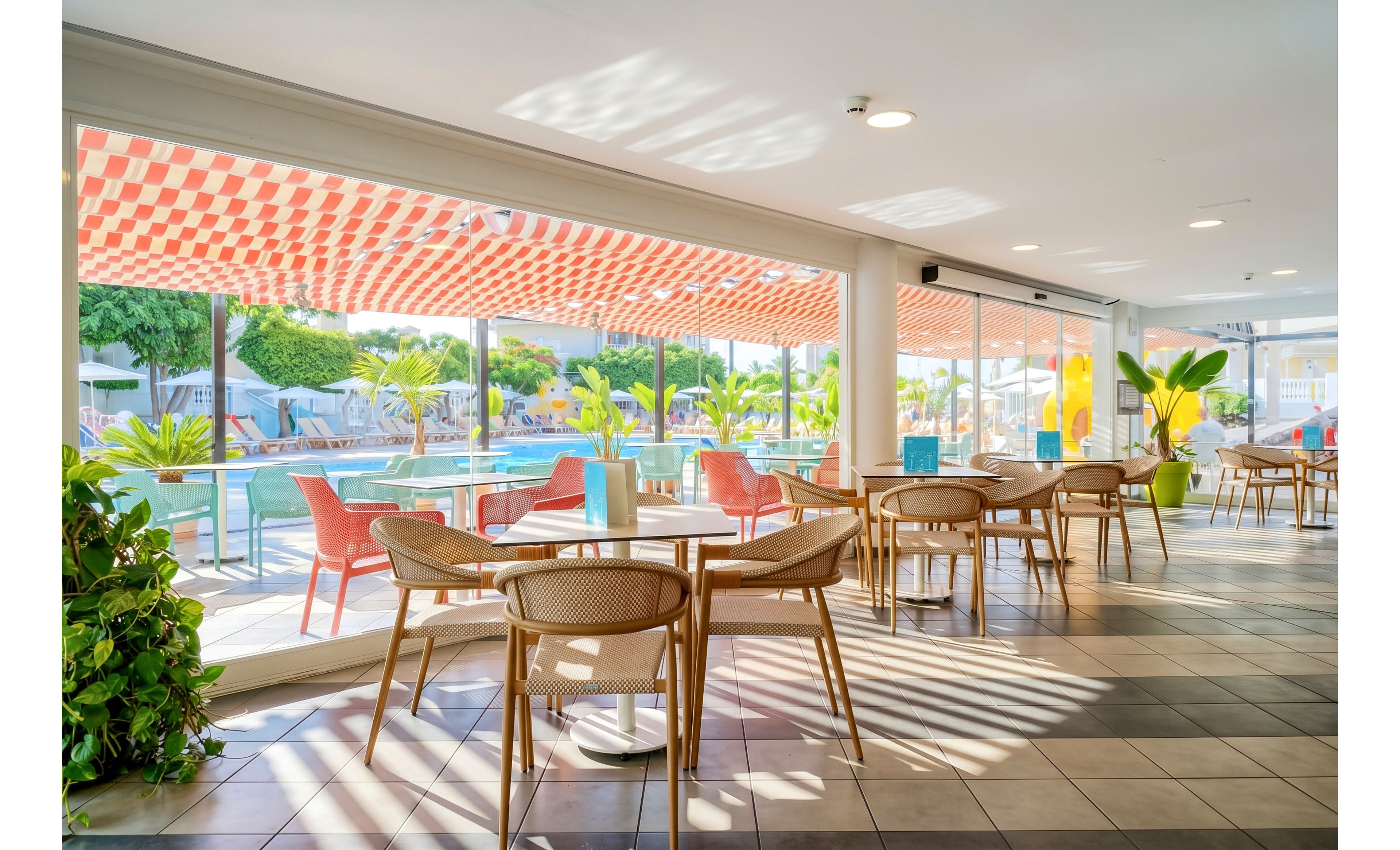 A modern poolside café features round tables and wicker-style chairs arranged beside large floor-to-ceiling windows. Outside, a swimming pool is surrounded by palm trees and sun loungers, creating a relaxed resort atmosphere. A red-and-white checkered awning casts patterned shadows across the tiled floor. Colorful accents, including turquoise menus and orange chairs, add a cheerful touch to the airy interior.