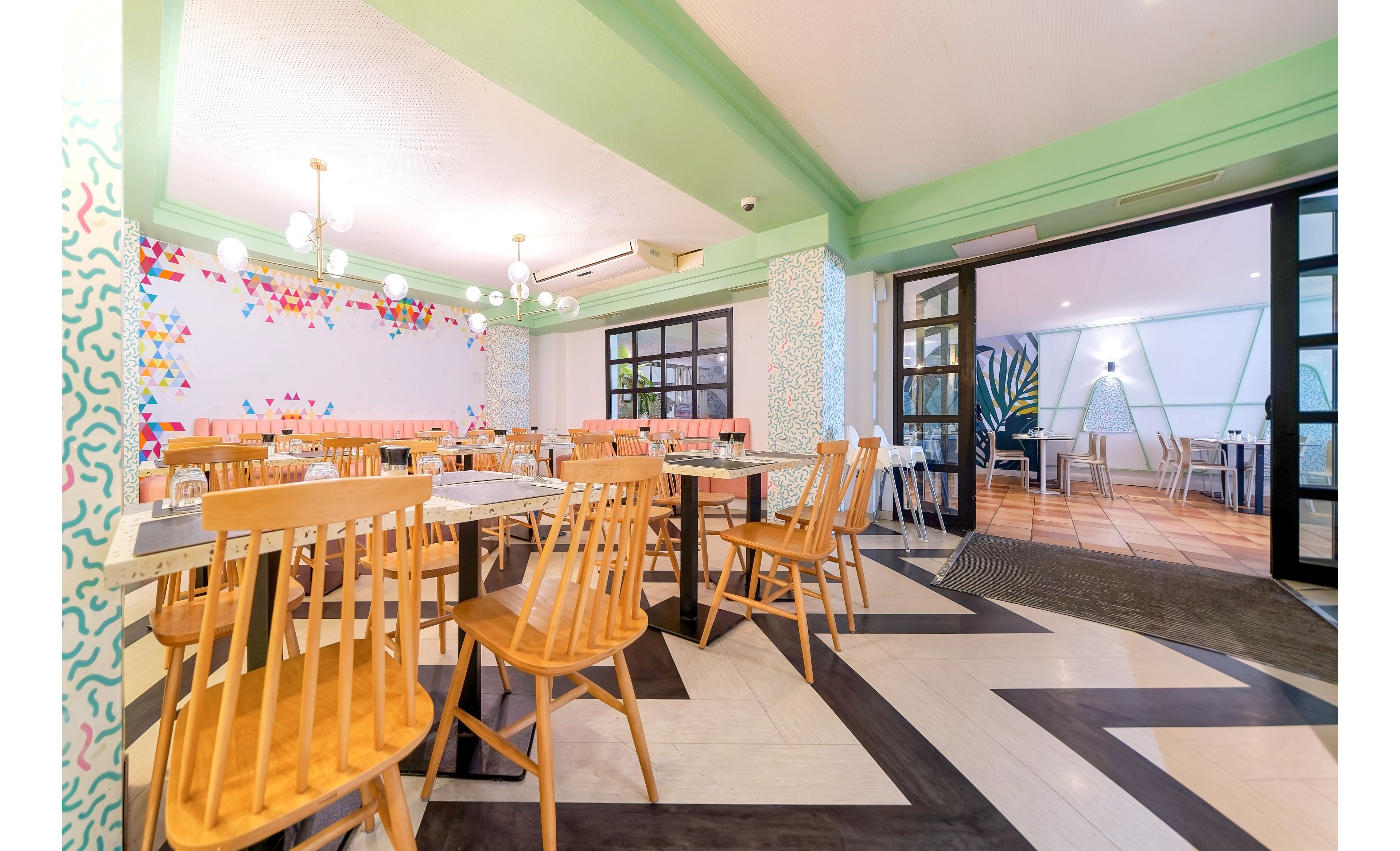 Contemporary restaurant dining area featuring light wooden chairs and tables arranged neatly across the room. The space has pastel green walls, geometric floor patterns, and colorful abstract artwork on one side. Pendant lights hang from the ceiling, creating a warm and inviting atmosphere. An open doorway leads to an adjacent seating area with additional tables and chairs, enhancing the airy, spacious feel.