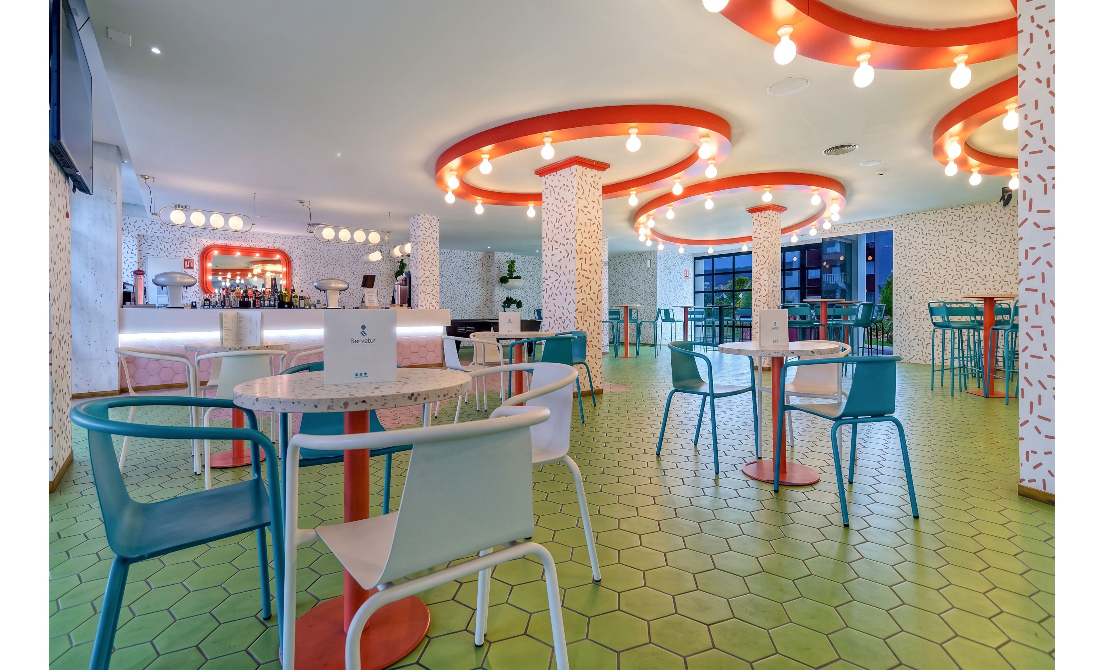 Bright contemporary cafe interior featuring round tables and mixed-color chairs on a green hexagonal tile floor. Curved ceiling elements with warm hanging lights create a playful, retro-inspired atmosphere. The bar area in the background is illuminated with circular light fixtures and surrounded by patterned columns. Large windows and glass doors provide natural light and a view of the outdoor seating area.