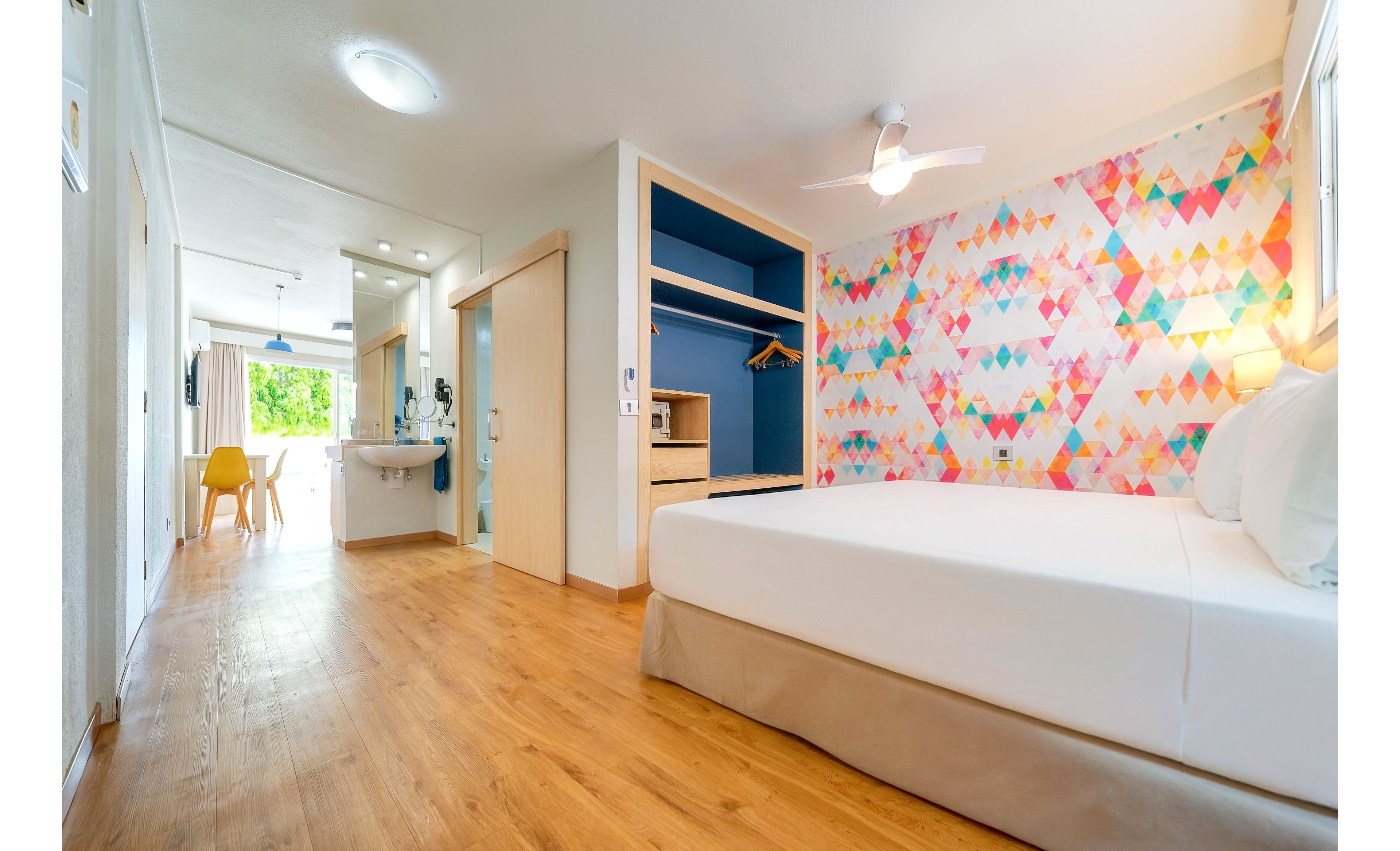 A spacious, modern hotel room features a large bed with white linens and a neutral bed base. One wall is covered with a vibrant geometric pattern in pink, orange, blue, and yellow tones, adding a playful touch to the space. The room has light wood flooring, an open wardrobe area, and a small vanity sink near the entrance. In the background, a sitting area with a table and chairs opens to a balcony with greenery and natural light.