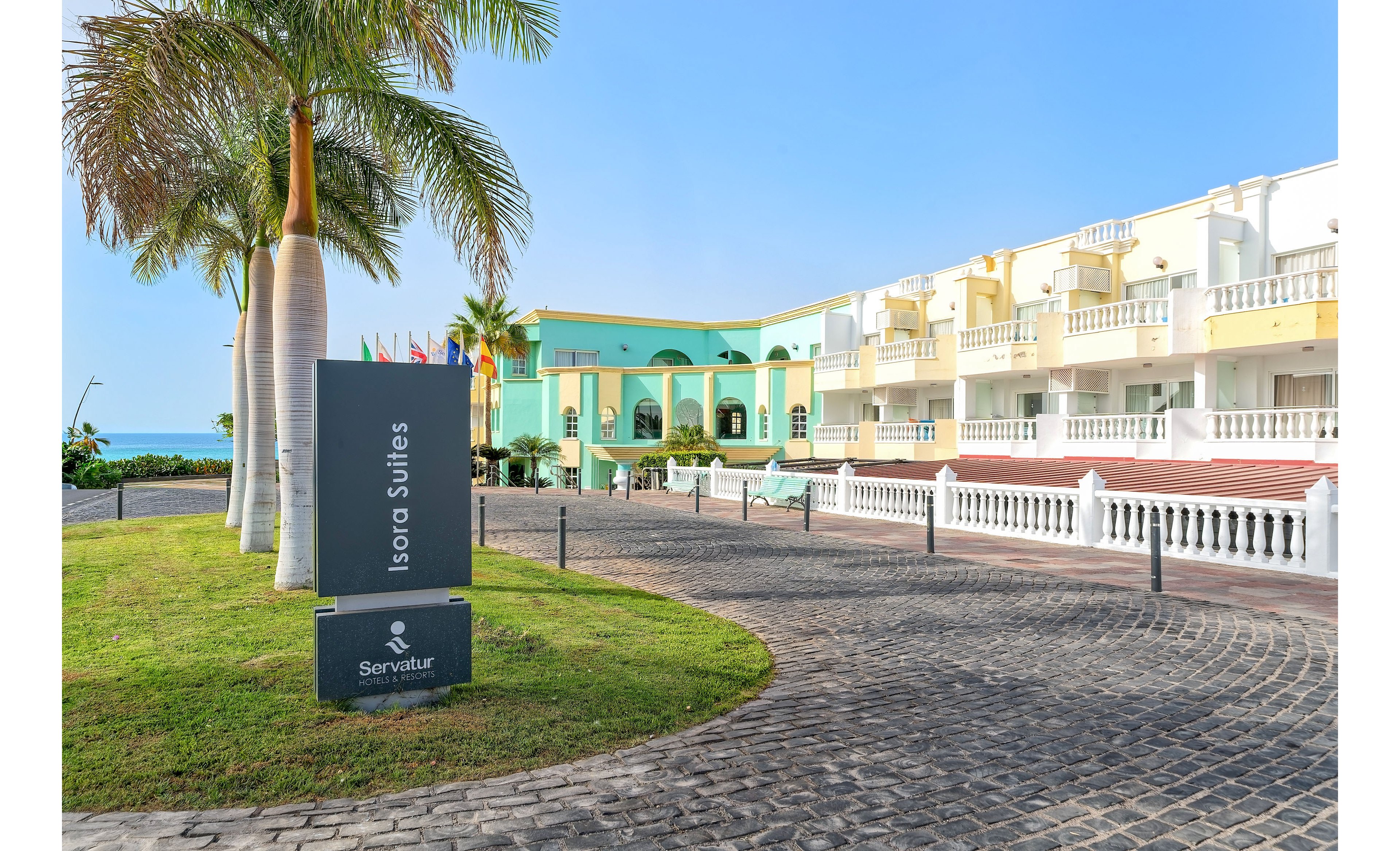 Exterior view of the Servatur Isora Suites hotel with pastel-colored buildings and balconies. A curved cobblestone driveway leads past palm trees and manicured lawns toward the entrance. The hotel sign reading "Isora Suites" and "Servatur" is prominently displayed in the foreground. Clear blue sky and a glimpse of the sea in the background create a bright, relaxed vacation atmosphere.