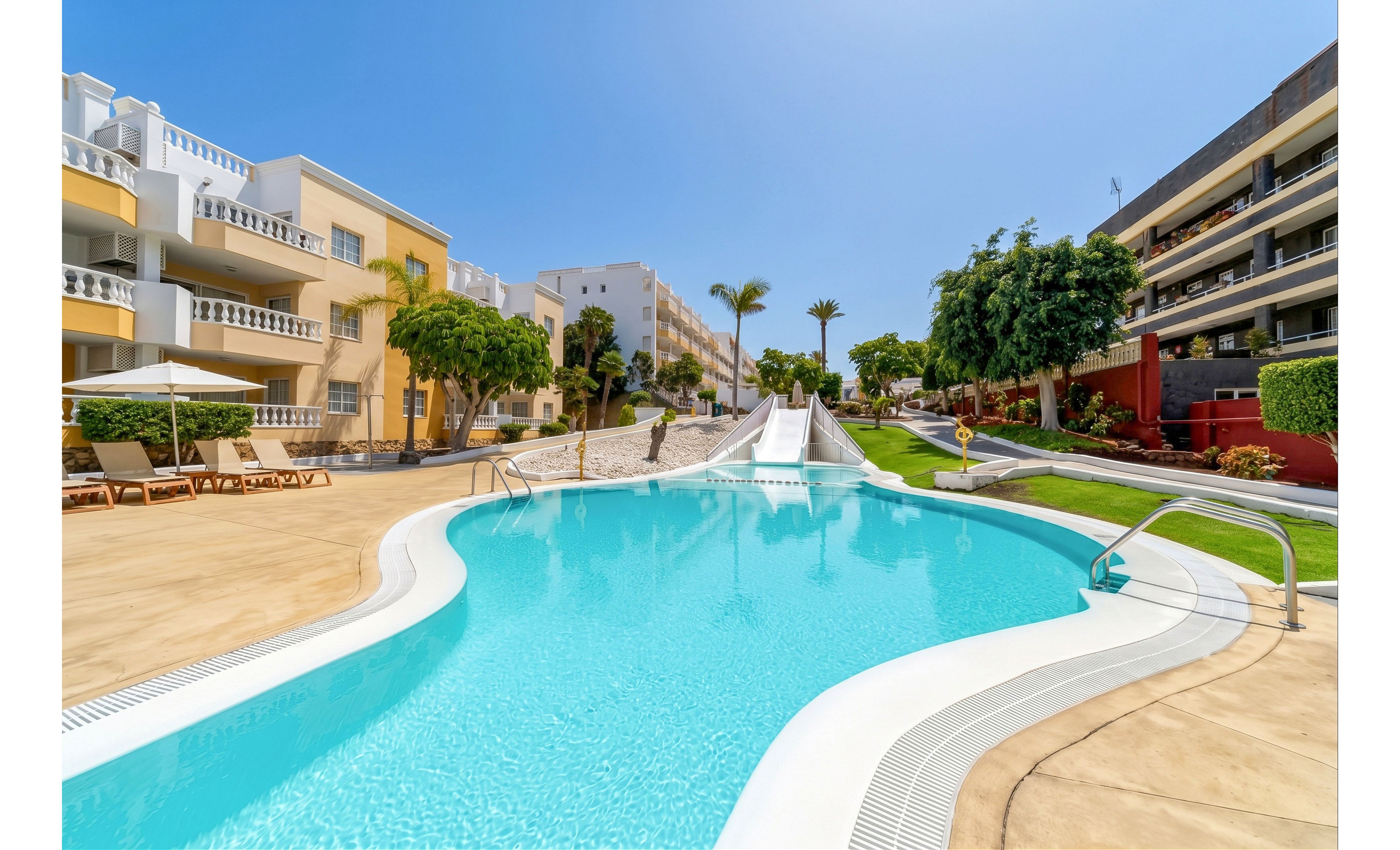 A bright outdoor swimming pool curves through a modern resort courtyard under a clear blue sky. Sun loungers and umbrellas line the poolside near multi-story apartment-style buildings. A central waterslide descends into the pool, framed by palm trees and manicured lawns. The scene conveys a relaxed vacation atmosphere with clean architecture and vibrant colors.