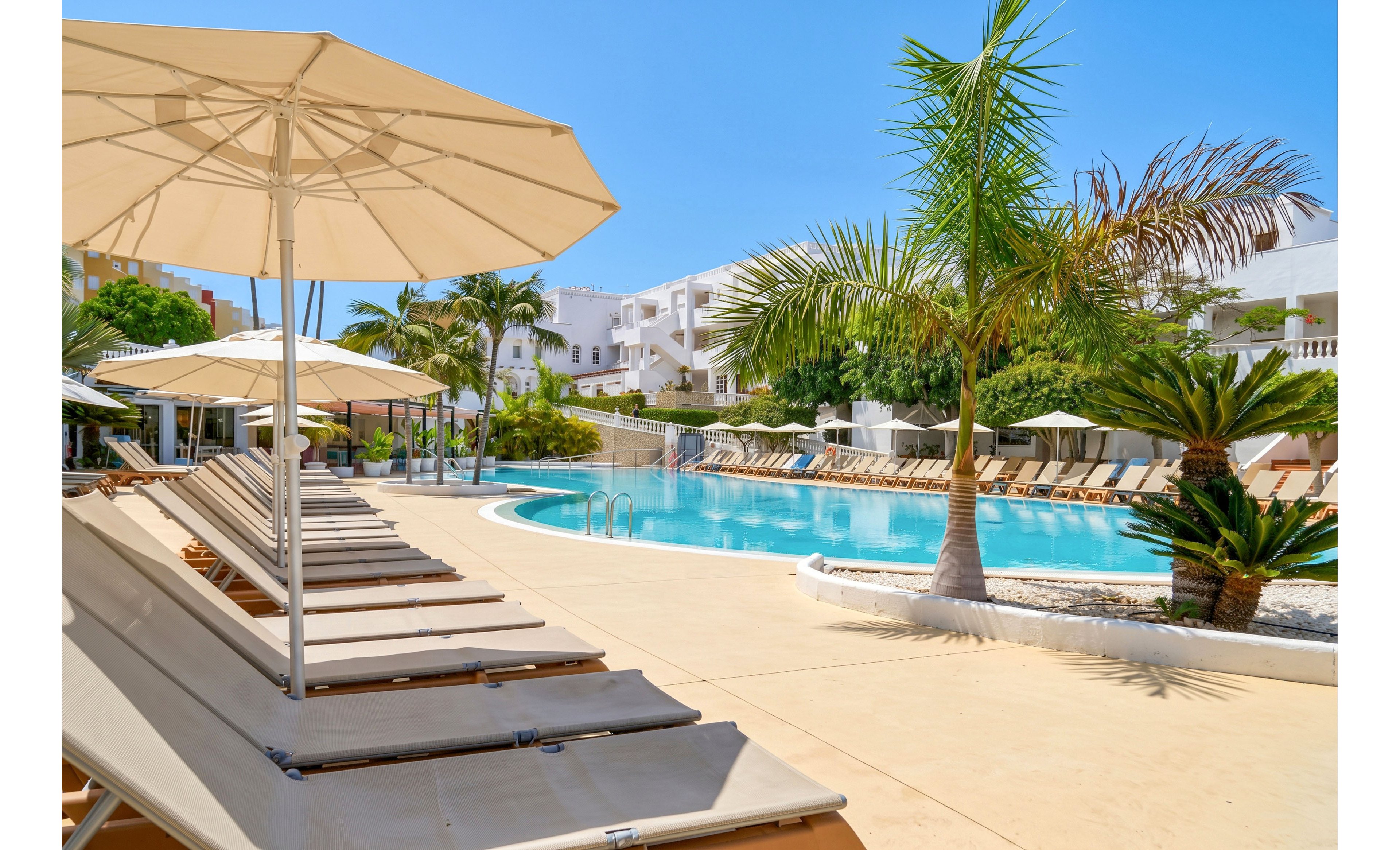 A bright resort swimming pool is surrounded by rows of beige sun loungers and large white parasols. Palm trees and lush greenery frame the pool area, adding a tropical feel to the scene. White modern buildings rise in the background under a clear blue sky. The overall atmosphere is calm, inviting, and ideal for a relaxing vacation day.