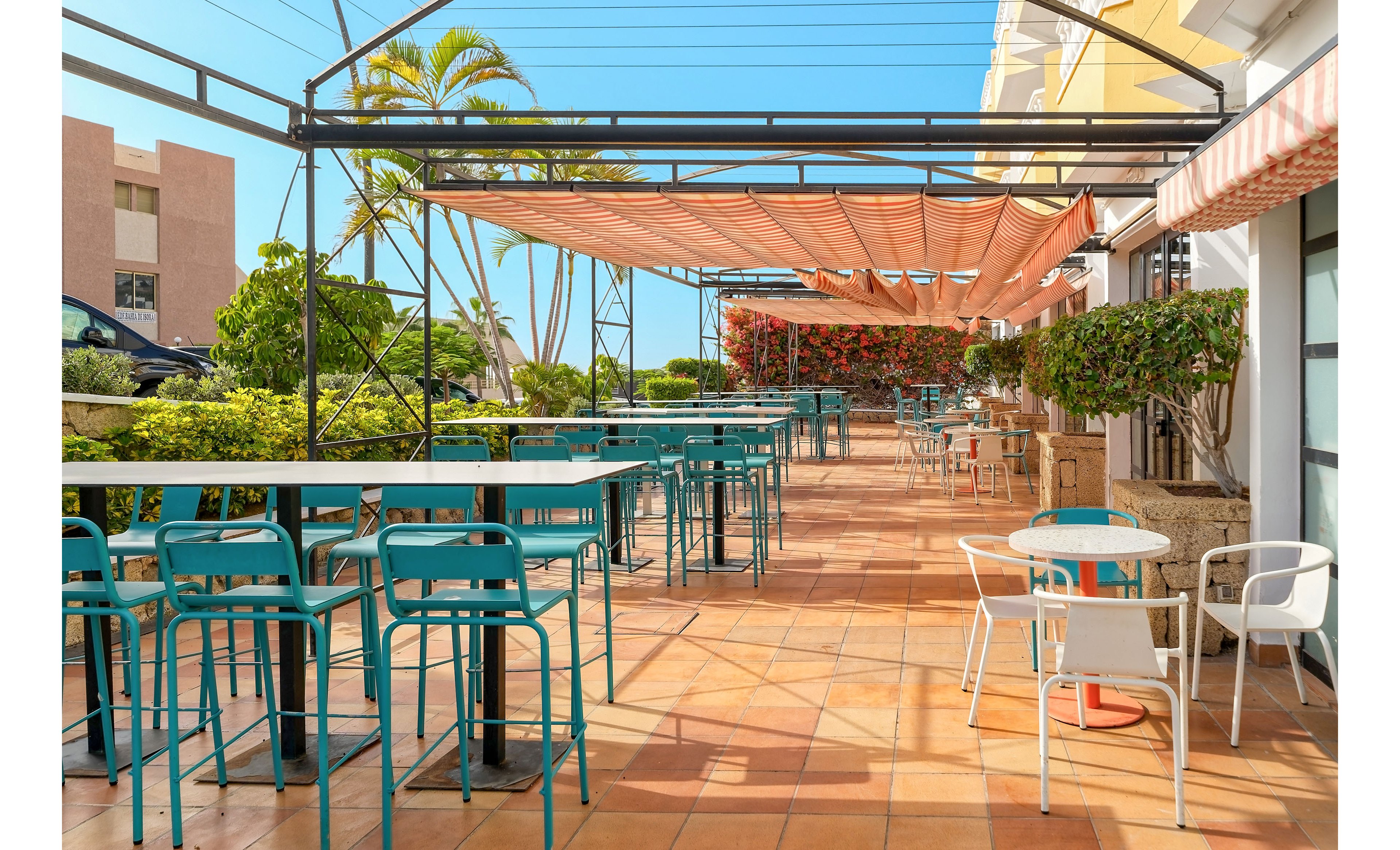 An empty outdoor cafe terrace features turquoise bar stools and white round tables arranged on a tiled floor. Orange fabric awnings provide shade above the seating area, while lush green shrubs and flowering plants line the edges. Palm trees and nearby buildings are visible in the background under a clear blue sky. The scene suggests a relaxed, warm-weather setting ideal for casual dining or drinks.
