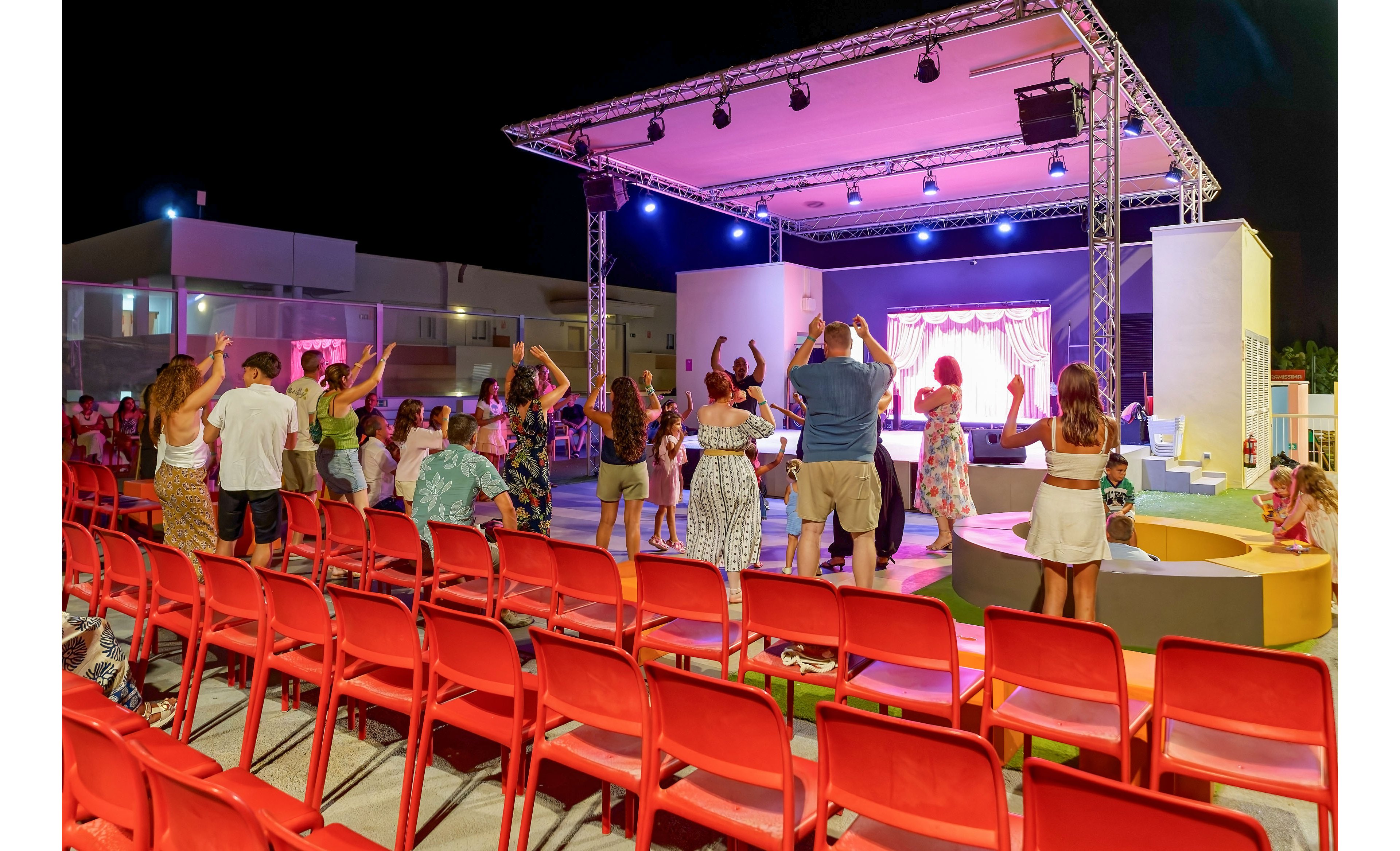 A group of people stand and dance in front of an outdoor stage at night. Bright colored lights illuminate the performance area, creating a festive atmosphere. Rows of empty red chairs are visible in the foreground, leading toward the crowd near the stage. The scene suggests a holiday resort or community event with families and friends gathered for entertainment.