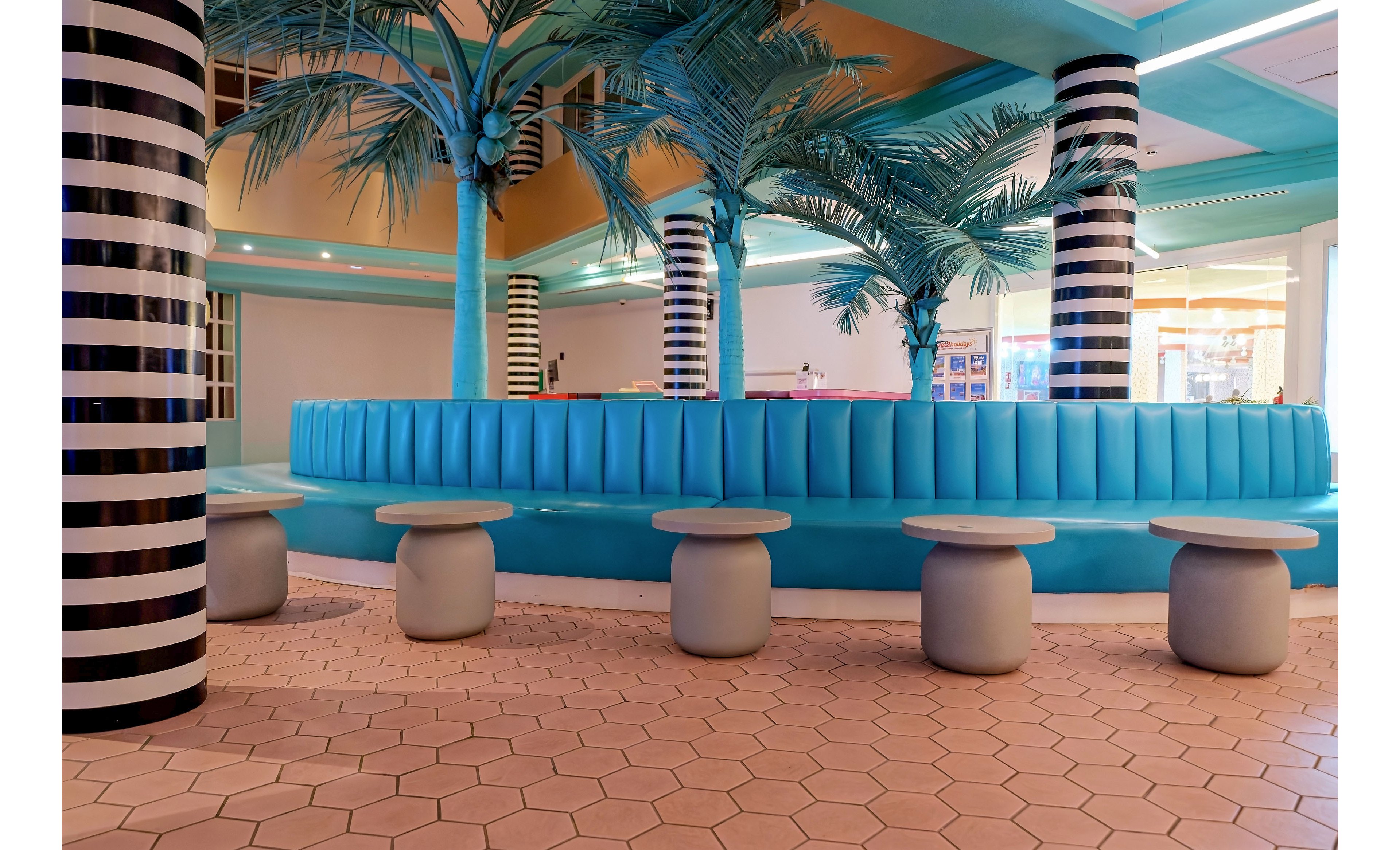 A contemporary indoor lounge features a curved turquoise banquette surrounded by cylindrical stools. Tall striped columns and artificial palm trees create a playful, tropical-inspired atmosphere. The floor is covered with light hexagonal tiles, and soft ambient lighting highlights the bold geometric design. The space appears empty, emphasizing the architectural details and vibrant color palette.