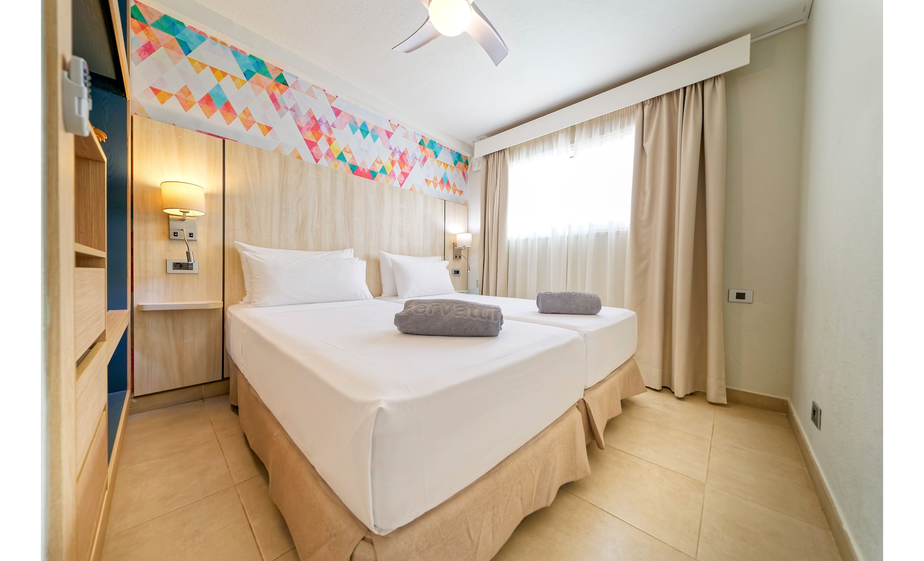 A clean, modern hotel room features two neatly made twin beds with white linens and folded gray towels on top. Light wood furniture and neutral beige flooring create a warm, minimalist atmosphere. A colorful geometric wall art strip adds a playful accent above the headboard area. Soft natural light filters through sheer curtains, complemented by a ceiling fan and bedside lamps for a comfortable stay.