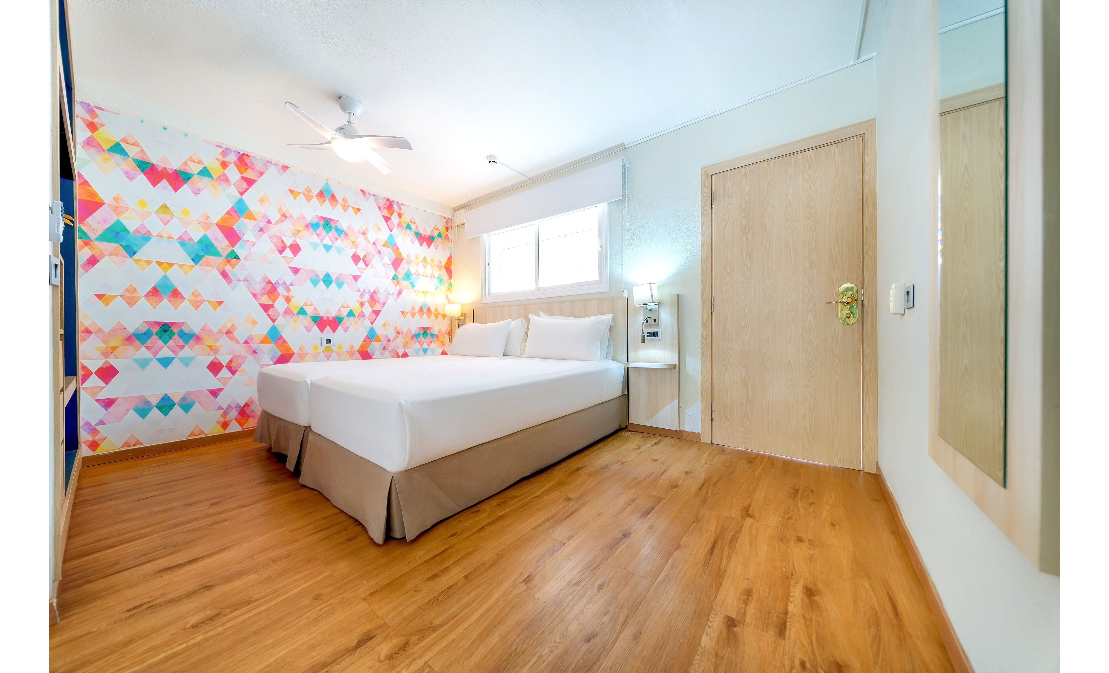 A contemporary hotel room features two neatly made twin beds with white linens and beige bed skirts. The space has warm wooden flooring and a light wooden door, creating a clean and inviting atmosphere. One wall is decorated with a colorful geometric pattern, adding a vibrant accent to the otherwise minimalist design. Natural light enters through the window above the beds, complemented by a ceiling fan and bedside lighting.