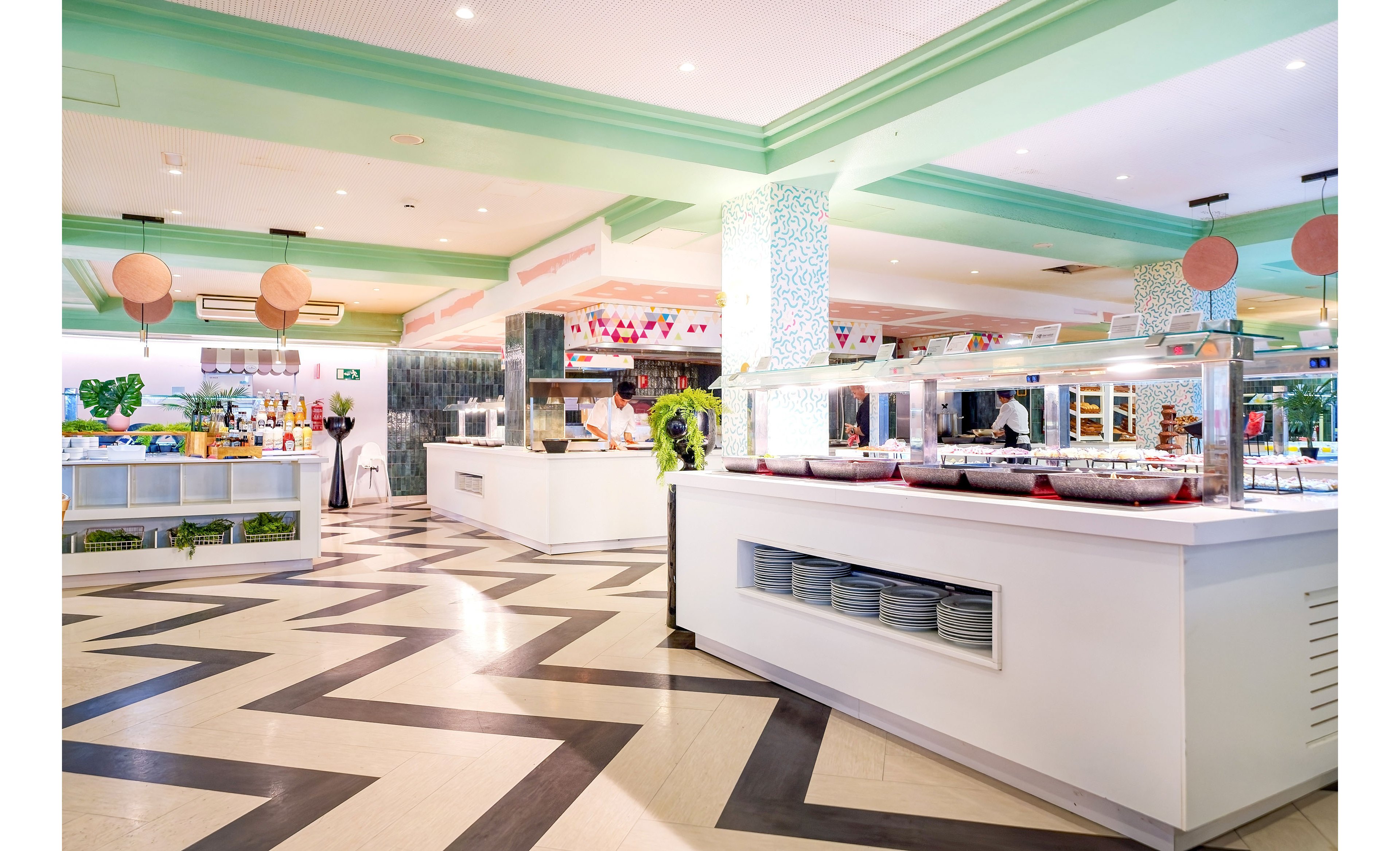 Spacious self-service buffet counters stand in a brightly lit restaurant interior. The room features zigzag patterned flooring, mint green ceiling accents, and white serving stations with covered food trays. Decorative hanging lanterns and plants add a fresh, contemporary atmosphere. The scene is clean, organized, and ready to serve guests in a casual dining setting.