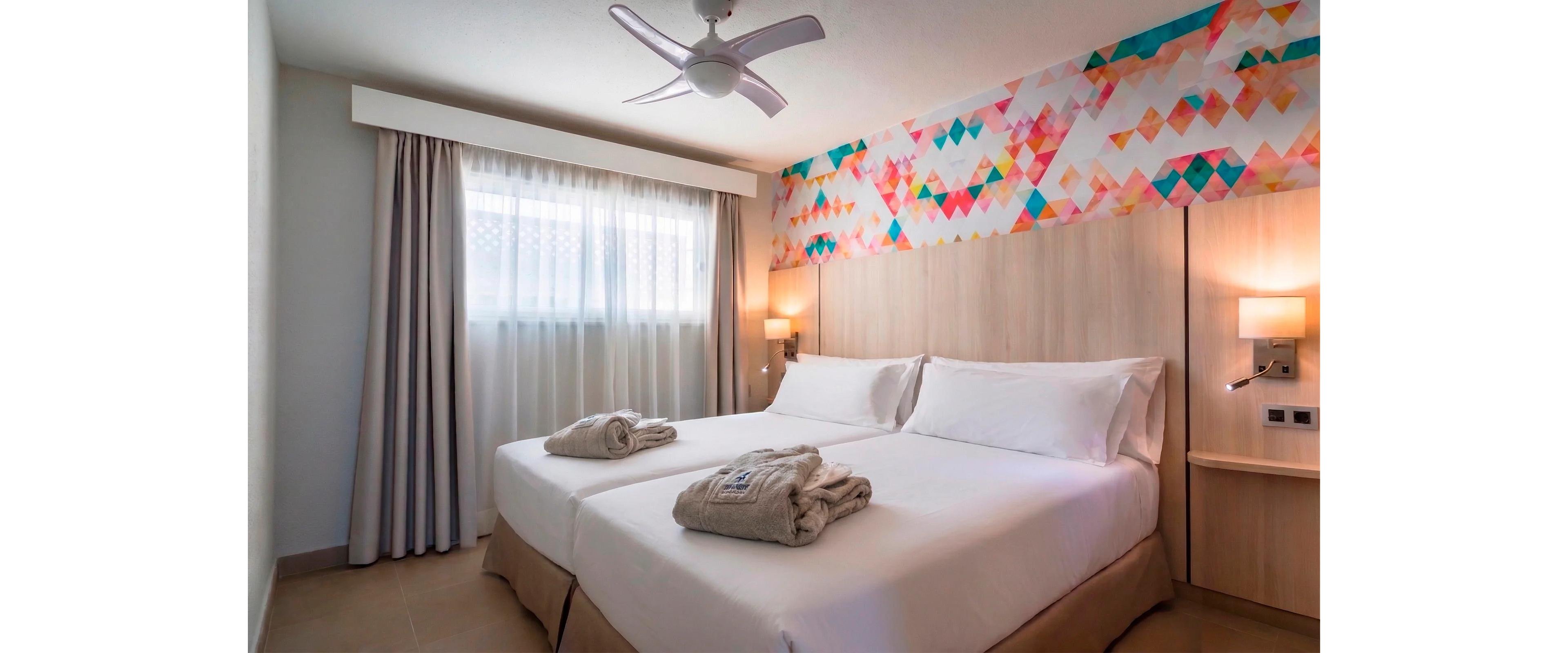 A contemporary hotel bedroom features two neatly made twin beds pushed together, each topped with folded towels. Soft natural light filters through sheer curtains covering a large window, creating a calm and inviting atmosphere. The room is decorated with light wood furniture and a colorful geometric accent strip running along the upper wall. Warm bedside lamps, a ceiling fan, and neutral tones enhance the cozy, minimalist design.