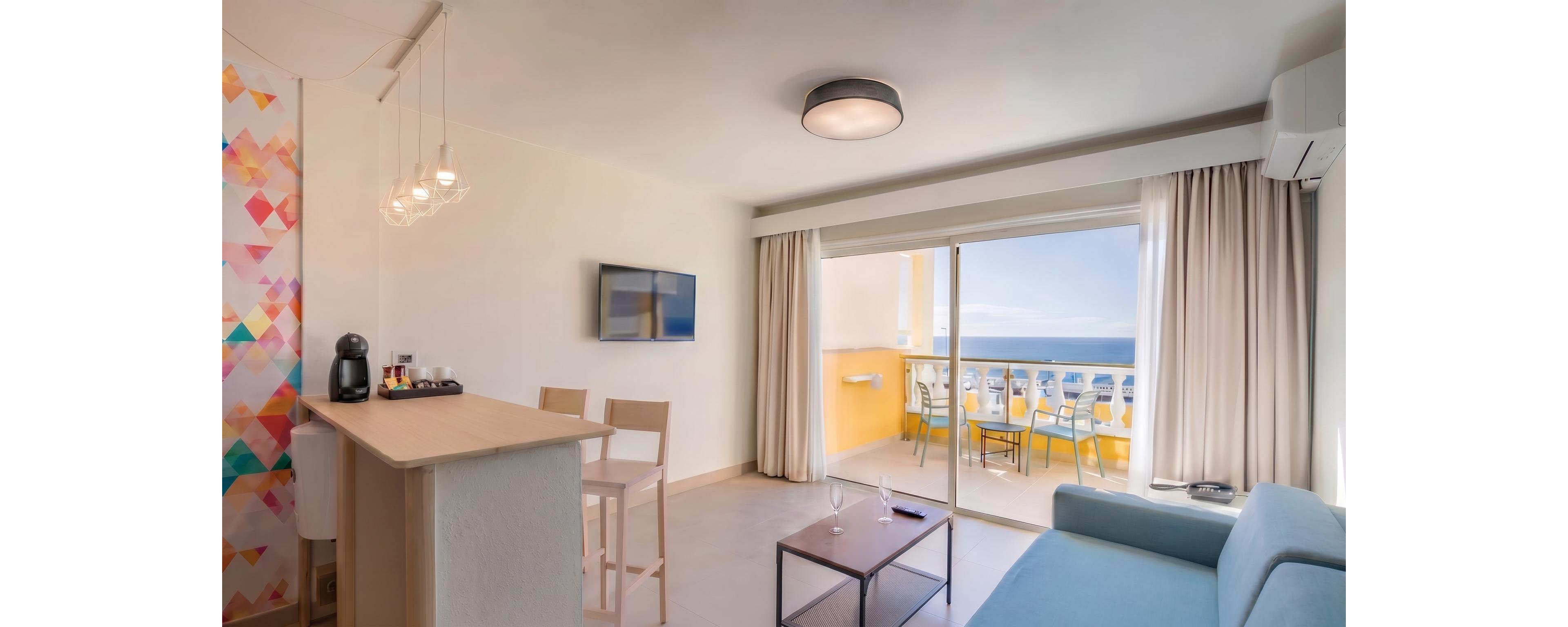 A bright modern apartment living room opens onto a spacious balcony overlooking the sea. The interior features a light blue sofa, a small coffee table with a wine bottle and glasses, and a wall-mounted television. A breakfast bar with stools and a coffee machine sits near a colorful geometric accent wall. Large sliding glass doors and neutral curtains allow natural light to fill the space, creating an airy coastal atmosphere.