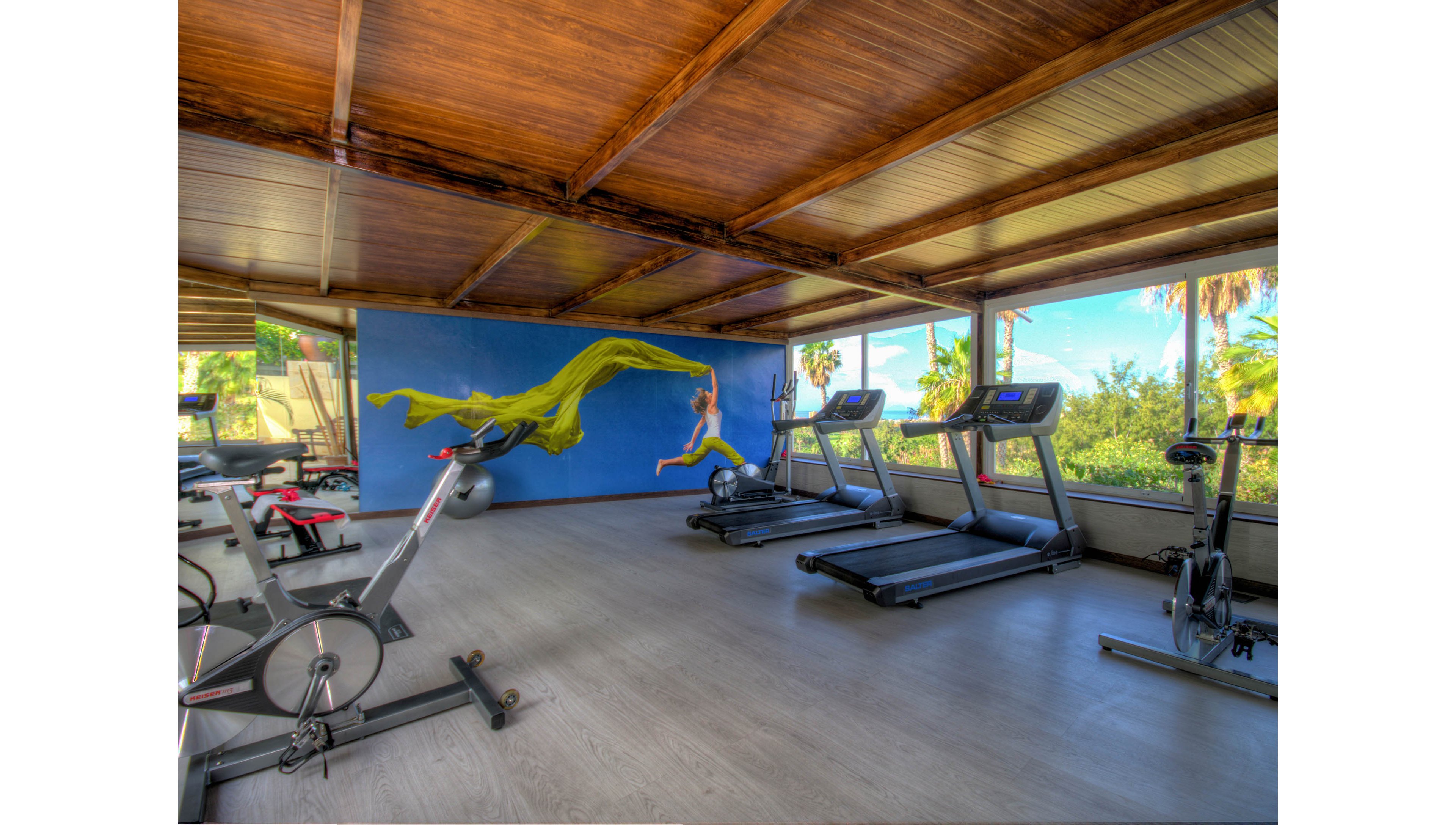A modern gym room features several treadmills and stationary bikes arranged along large windows. Natural light floods the space, highlighting the wooden ceiling and light-colored flooring. A vibrant wall mural of a running figure with a flowing yellow fabric adds energy and color to the interior. Lush greenery is visible outside, creating an open and refreshing workout environment.