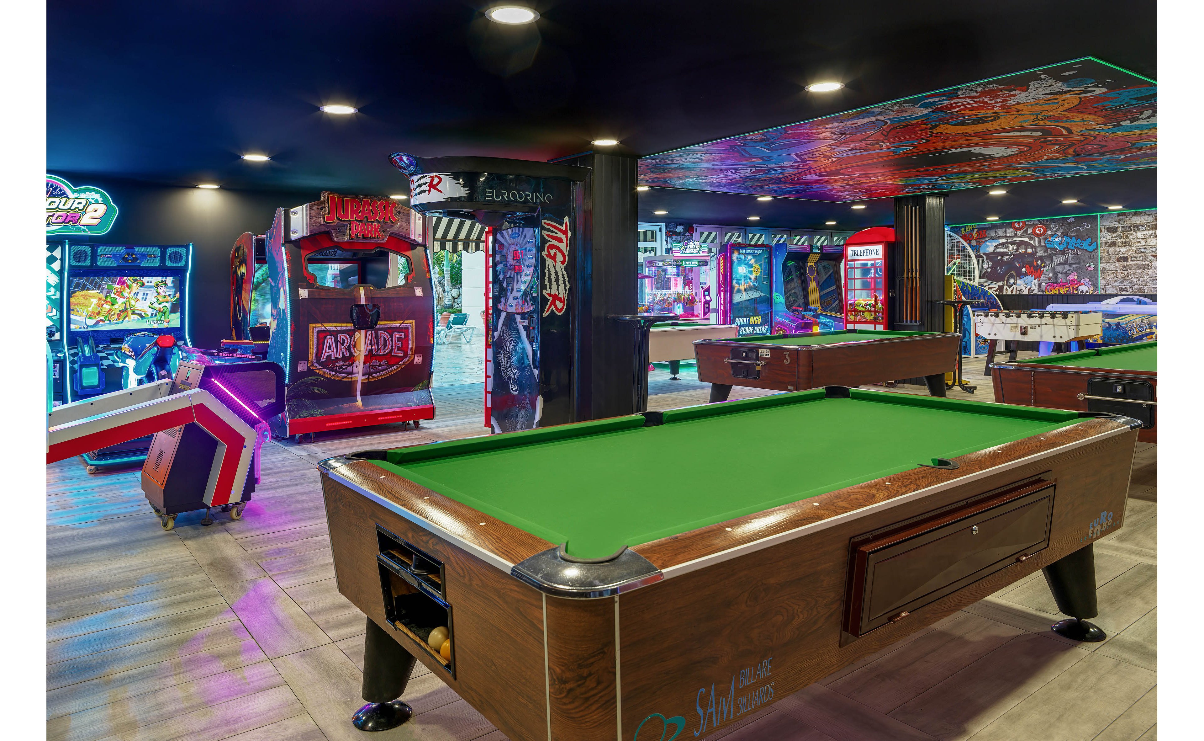 A brightly lit game room features a green-felt pool table in the foreground. In the background, various arcade machines, including a large basketball game and racing cabinets, create a colorful and dynamic atmosphere. Neon lights and illuminated screens add vibrant hues of blue, red, and purple throughout the space. The setting suggests an entertainment venue designed for leisure, fun, and social gaming.