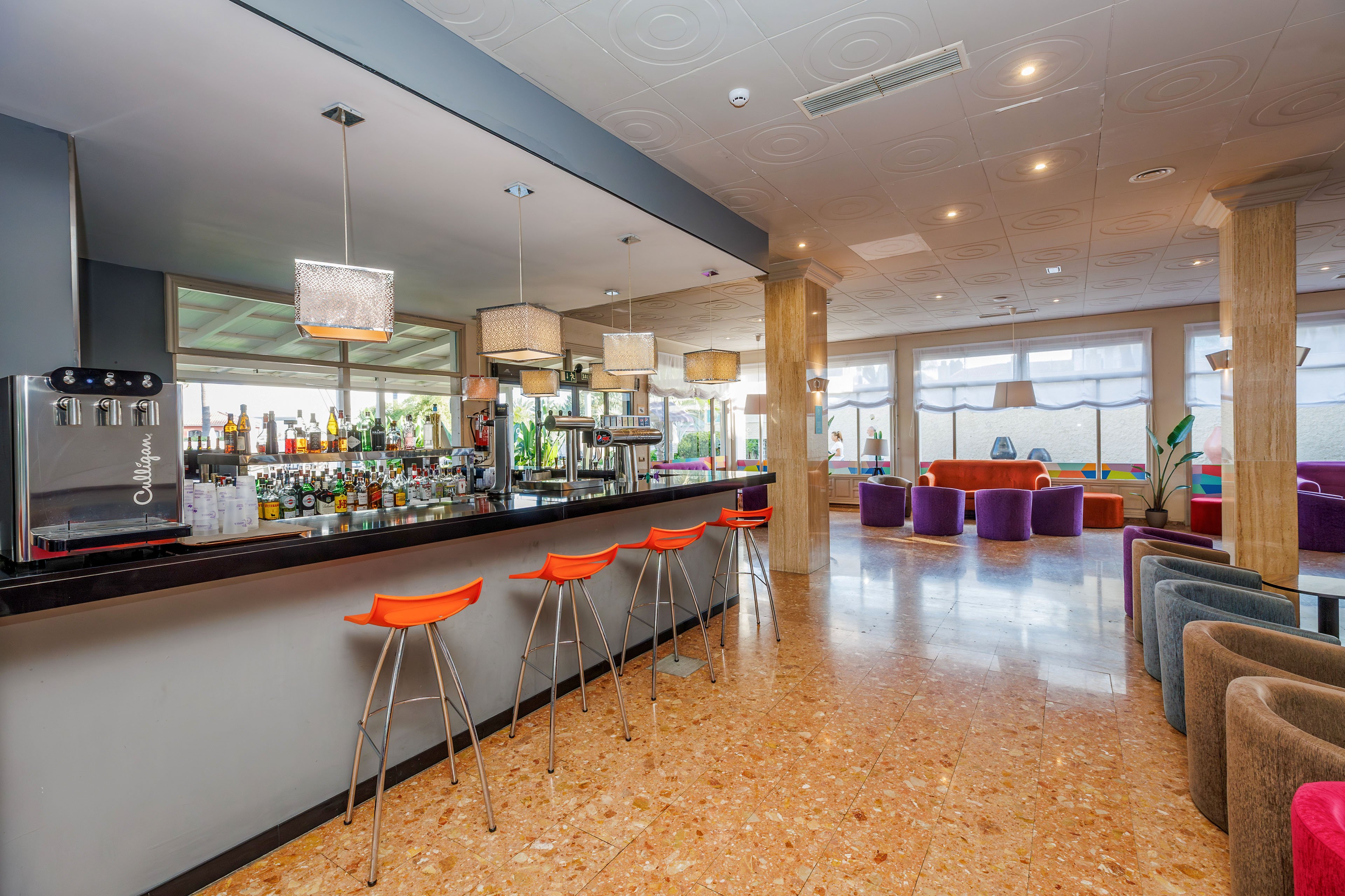 A contemporary hotel bar interior features a long counter with bright orange bar stools and a well-stocked selection of bottles. Pendant lights hang above the bar, illuminating the polished surface and stainless steel drink dispensers. In the background, a spacious lounge area includes colorful armchairs and sofas arranged around low tables. Large windows and glass doors bring in natural light, highlighting the terrazzo-style floor and neutral-toned columns.