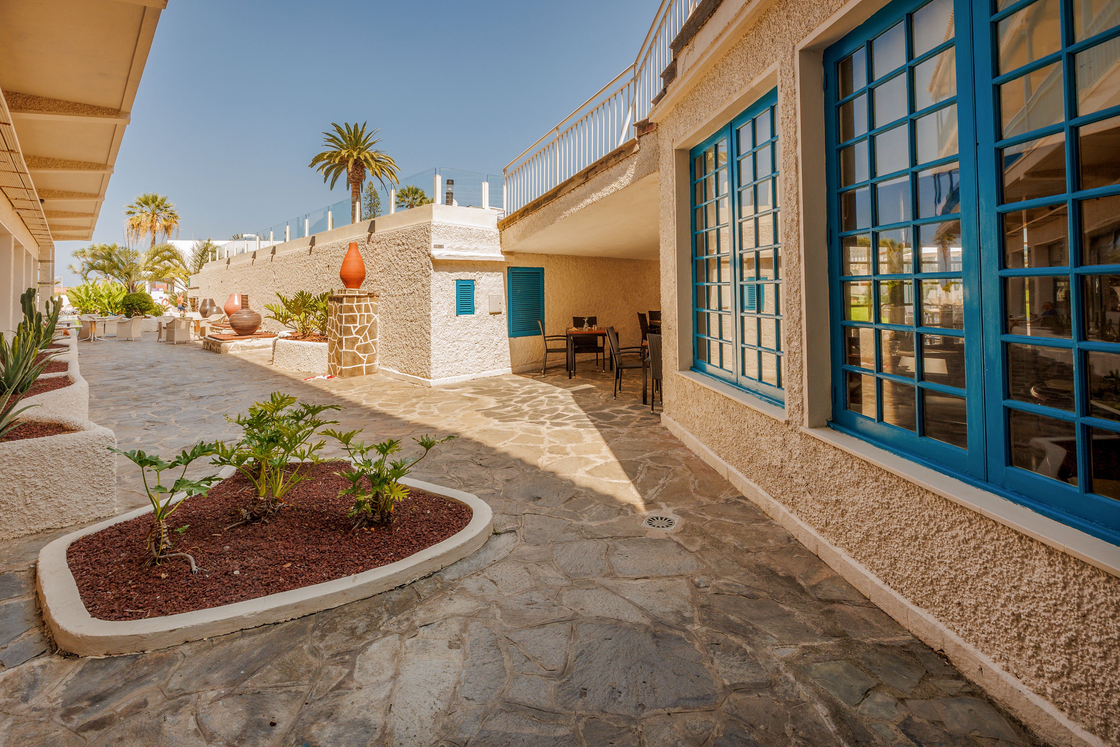 A bright stone courtyard stretches between white stucco buildings with vivid blue-framed windows and doors. Potted plants and small landscaped beds with red mulch add greenery along the walkway. Outdoor tables and chairs sit in the shade beneath an overhang, suggesting a relaxed seating area. Palm trees and a clear blue sky in the background enhance the warm, coastal atmosphere.